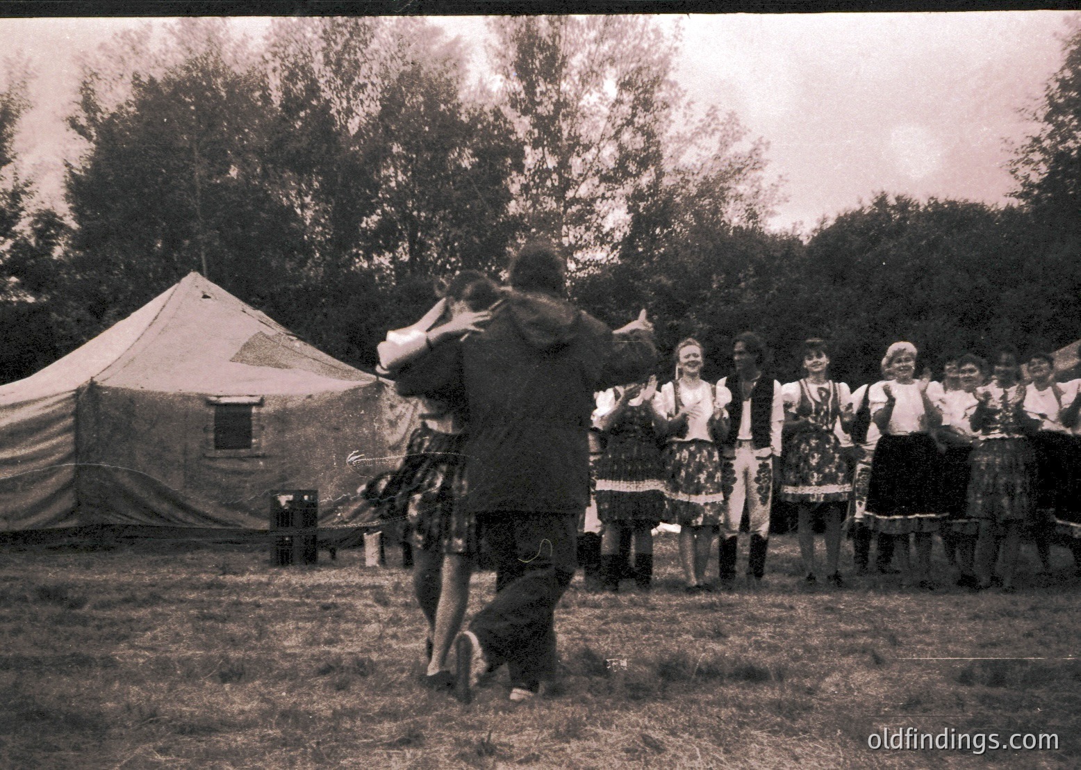 Vintage black-and-white photo of a traditional folk dance performance in an outdoor setting, likely Eastern Europe. Central male dancer in kilt-style attire leads a group of women in colorful embroidered dresses and headscarves. Tent and trees suggest a rural or festival venue, mid-20th century.