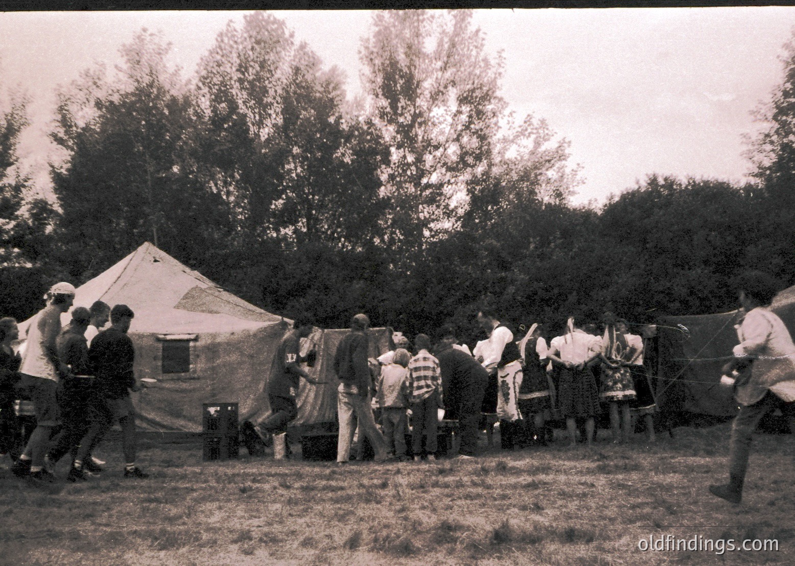 Group of children and adults in outdoor camping scene, likely 1950s–1970s. Tent with "13" marked on its side suggests organized youth activity. Mid-century clothing (plaid shirts, caps) and natural setting indicate a scout or summer camp gathering.