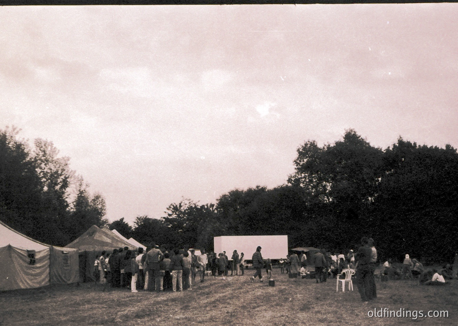 Vintage outdoor gathering in a rural setting, likely mid-20th century. A large screen displays content to an audience seated on wooden benches and standing. White tents and trees frame the scene, suggesting a temporary event or festival. Monochrome aesthetic enhances nostalgic atmosphere.