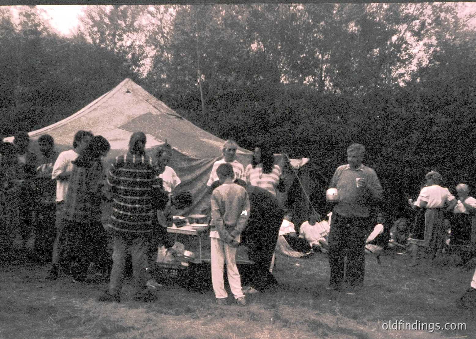 Vintage outdoor gathering with a group of people around a makeshift grill under a large canvas tent. Mid-20th century clothing suggests or . Wooded, rural setting with natural lighting. Ideal for historical research on camping or social events.