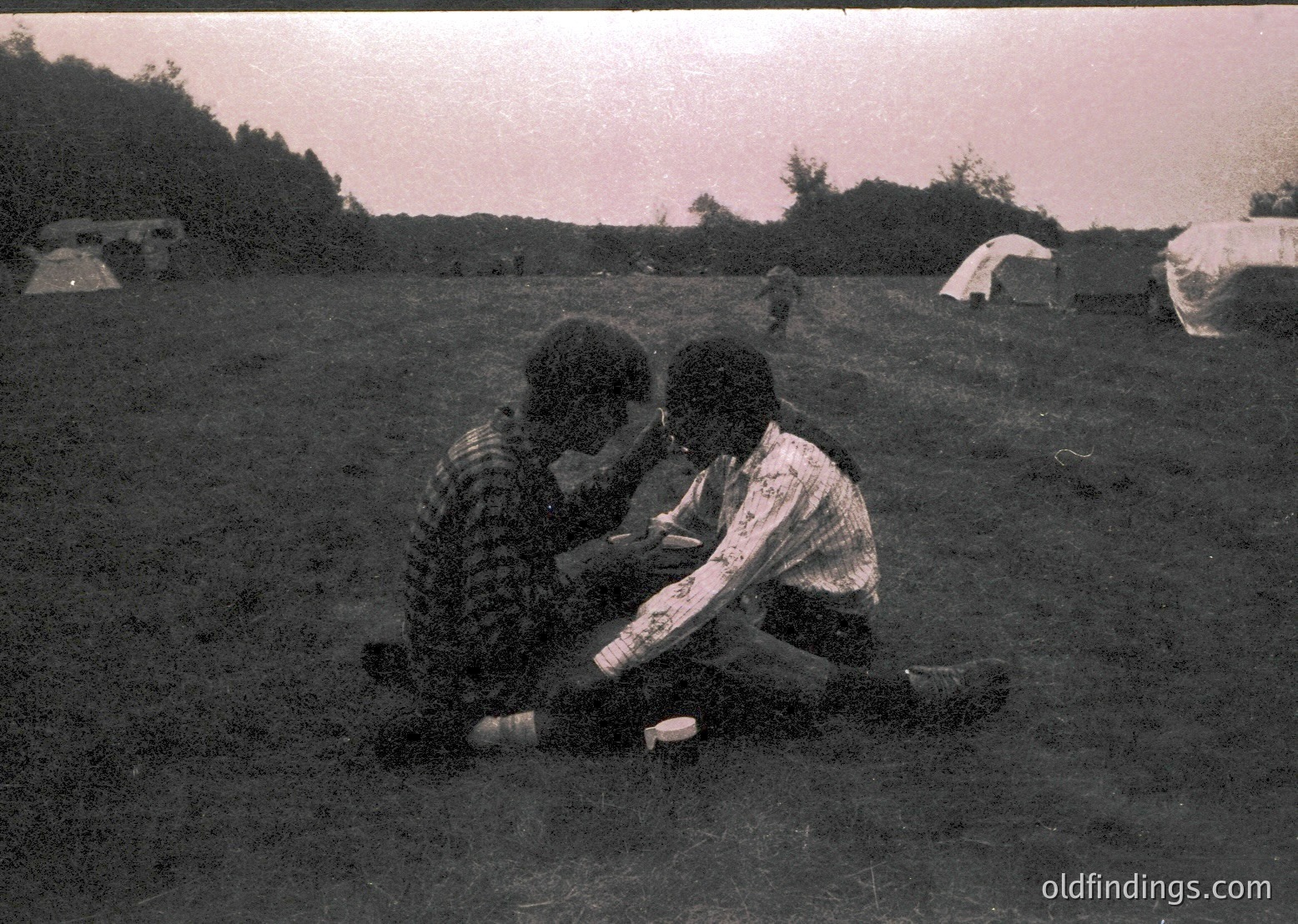 Vintage black-and-white photo of two individuals sitting closely in a grassy field, possibly mid-20th century. One wears a patterned sweater, the other a striped shirt. Tent structures and tents in background suggest camping or outdoor gathering. Soft focus and sepia tone enhance nostalgic atmosphere.