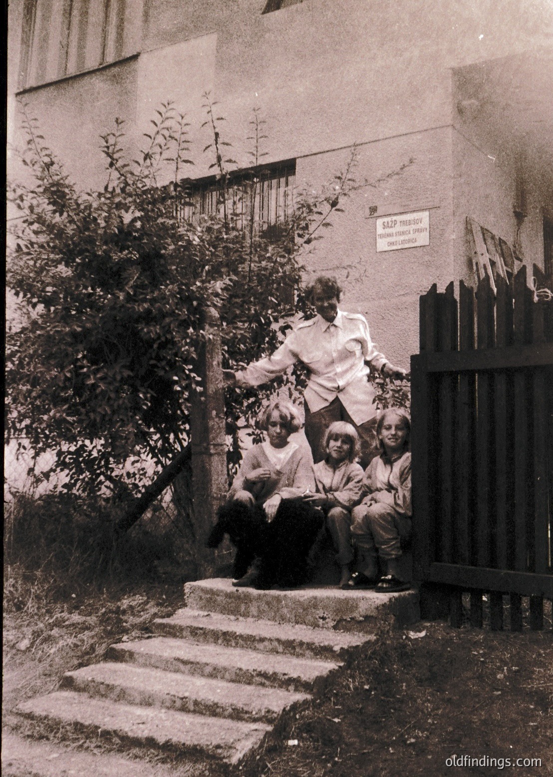 A black-and-white photo of a family posing on concrete steps beside a residential building, likely from the 1960s–1970s. The adult stands on the top step, while three children sit below, dressed in casual 20th-century attire. A sign on the building reads "САДОВОДСТВО" (horticulture) in Cyrillic, suggesting a Soviet-era community garden or housing complex. The setting appears to be a Soviet bloc country.