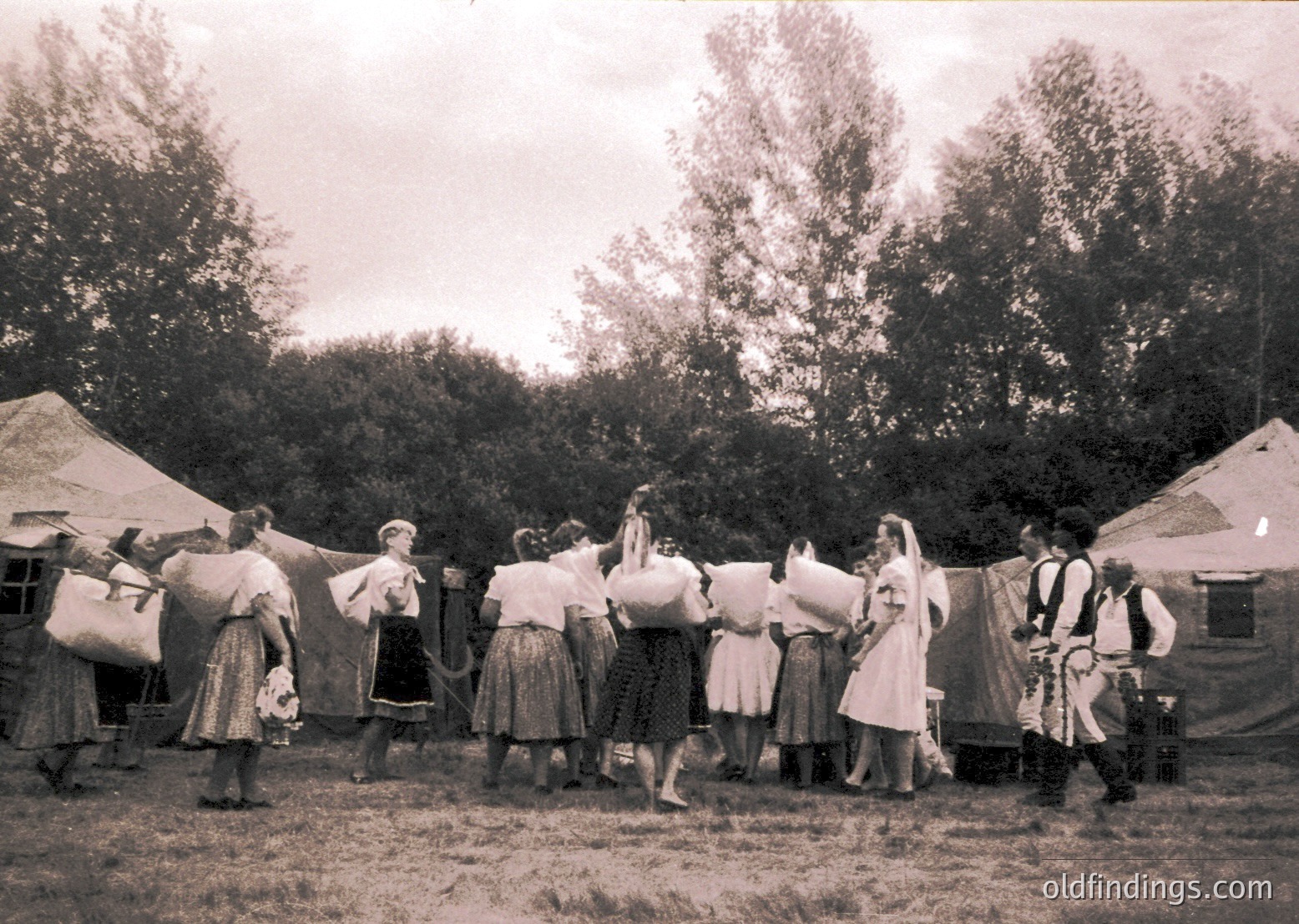 Vintage sepia-toned photo of a folk dance circle, likely Eastern European . Women in traditional skirts and blouses, men in embroidered vests, performing outdoors near canvas tents. Rural festival or cultural event setting.