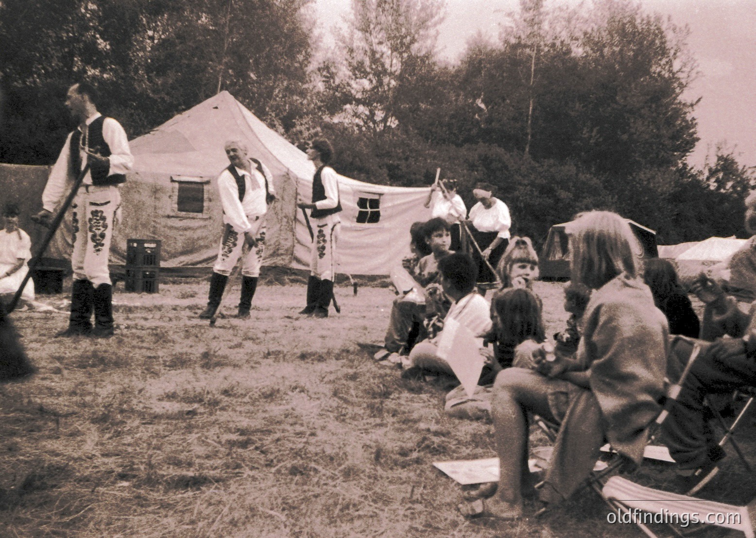 Reenactment scene featuring medieval-style performers in traditional attire—tunics, belts, and boots—engaging an audience seated on hay. Tent backdrop suggests an outdoor festival or historical camp. Black-and-white filter enhances vintage aesthetic.