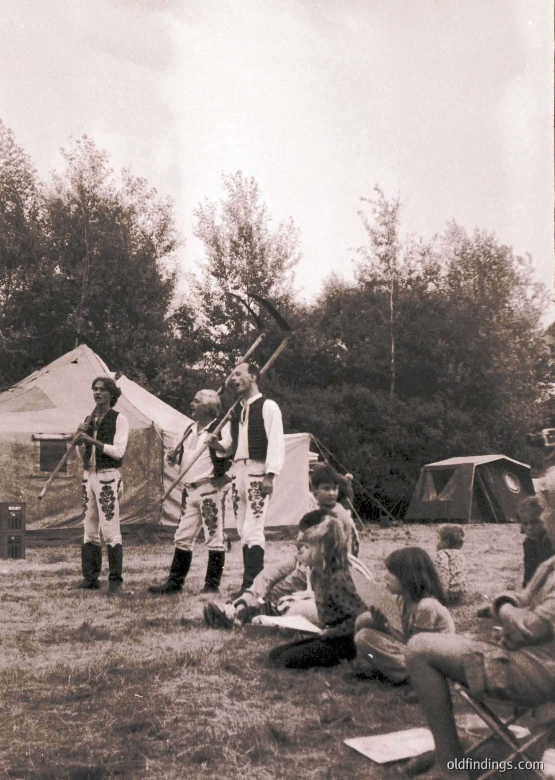 Vintage outdoor reenactment scene featuring three men in traditional Balkan military attire (19th-century style) with rifles and sabers. Tent camp and seated spectators in grassy area suggest historical festival or educational demonstration.