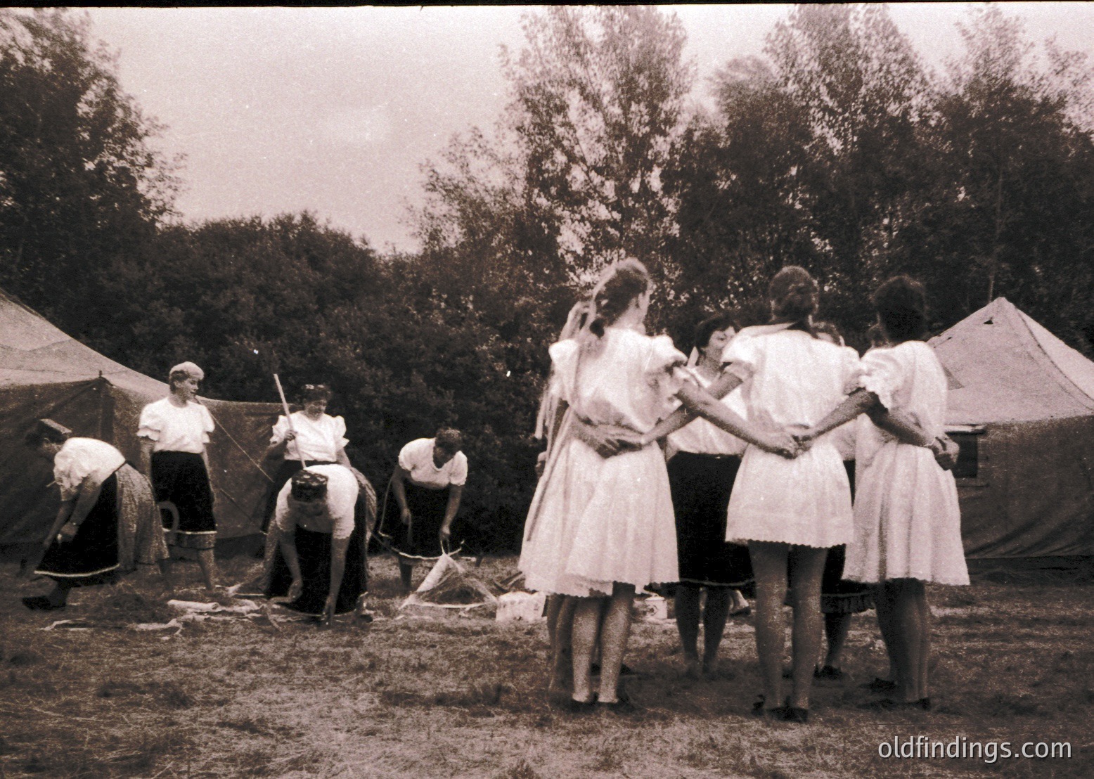Mid-20th century outdoor gathering with men in rolled-sleeve shirts and women in knee-length dresses. Group stands near a makeshift fire pit; one man shovels dirt while others observe. Tent and trees suggest a rural or camping setting. Likely 1950s–1960s.