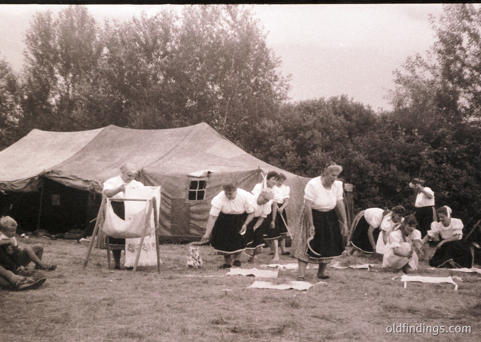 Vintage outdoor gathering featuring women engaged in traditional textile work under a large canvas tent. Folk costumes with embroidered aprons and headscarves suggest Eastern European cultural heritage. Mid-20th century (1940s–1960s) rural setting.