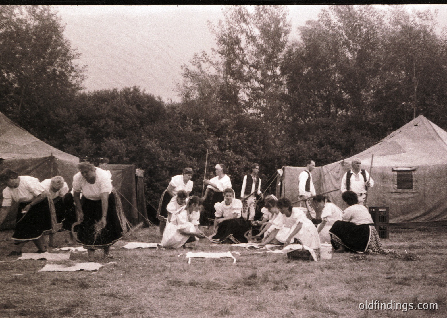 Vintage group engaged in traditional rope-making outdoors, seated on grass under large tents. Clothing suggests early-to-mid 20th century rural craftsmanship.