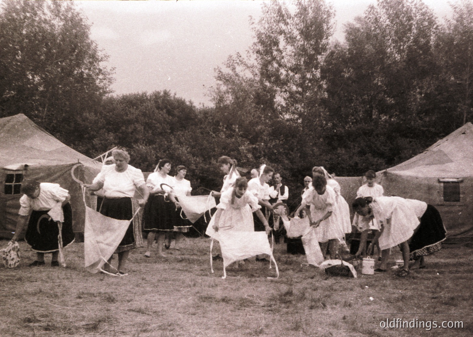 Vintage outdoor scene featuring a group of women in early 20th-century clothing, likely 1910s–1920s, performing a traditional folk dance. Women wear long skirts, aprons, and headscarves; one woman leads with a handkerchief or fan. Tents and trees suggest a rural or festival setting.