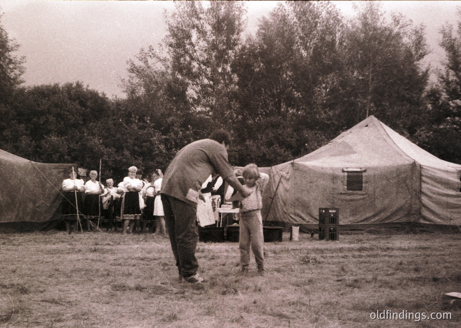 Military-style canvas tents in a grassy outdoor setting, mid-20th century. A man in a dark uniform adjusts a child’s cap, while others observe. Uniforms and tents suggest training or community event, likely Eastern Bloc era.