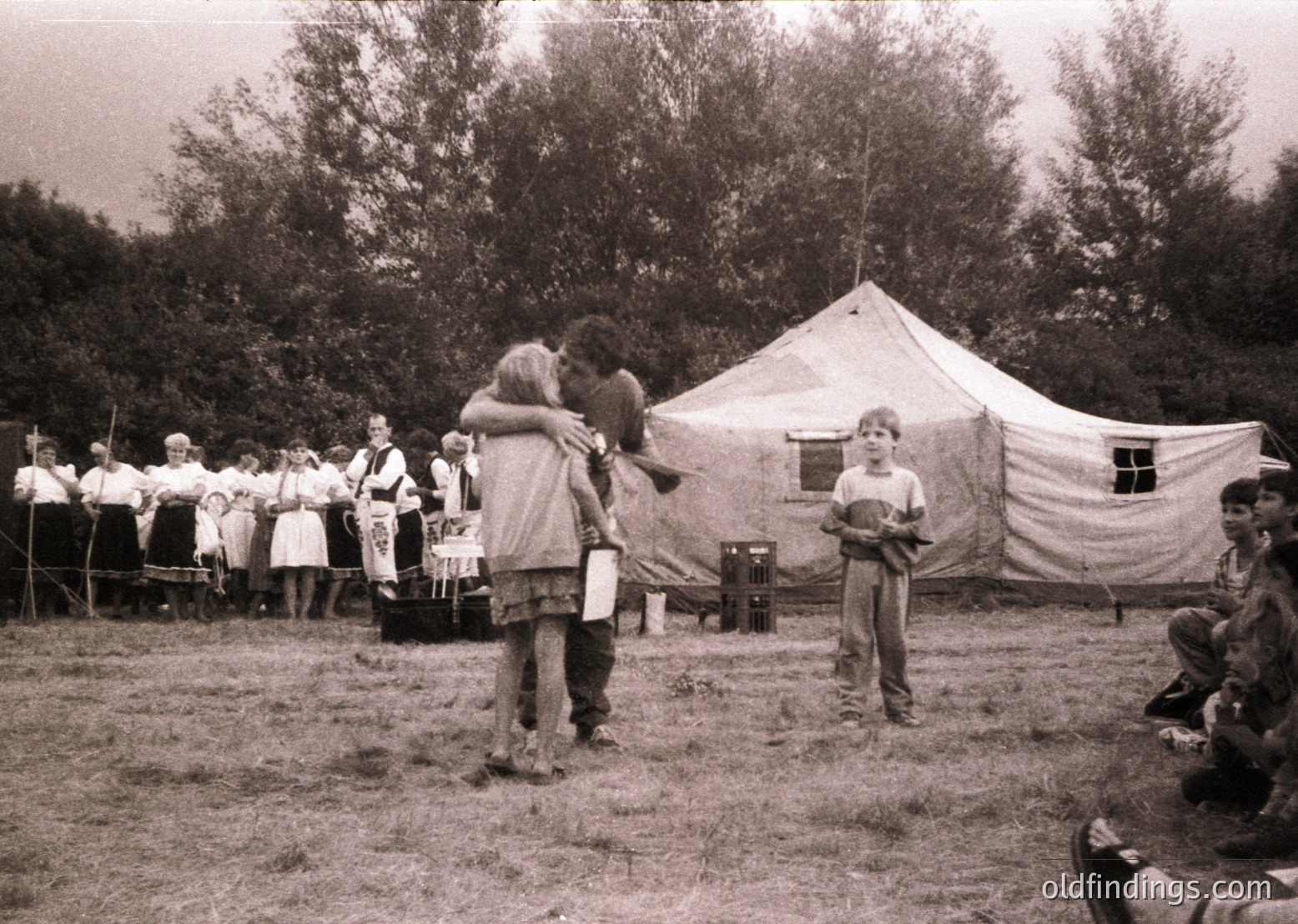 Mid-20th century outdoor gathering featuring a large canvas tent and group of people in casual attire. Central figures embrace, while a boy in shorts holds a small object. Background includes seated spectators and a forested area. Likely a festival or community event.