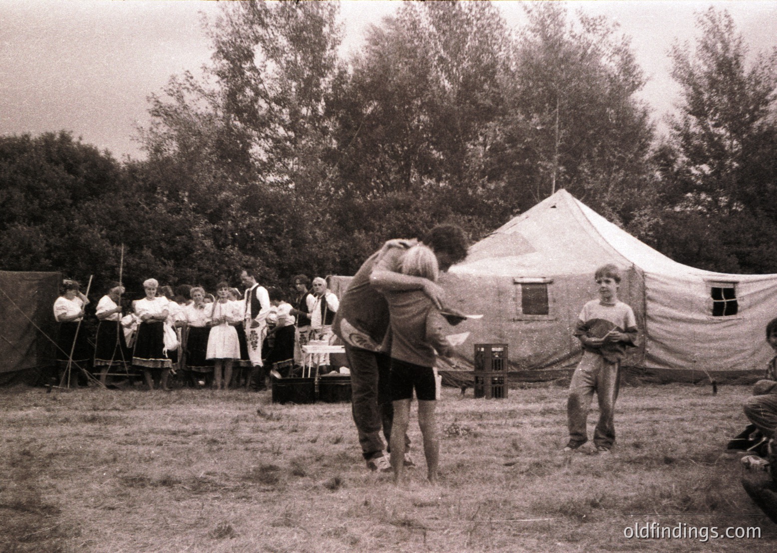 Mid-20th century outdoor gathering at a campsite or festival, featuring a large canvas tent and wooden structures. A couple dances while others observe, suggesting a communal or celebratory event. Clothing styles and tent design hint at 1950s–1960s era.