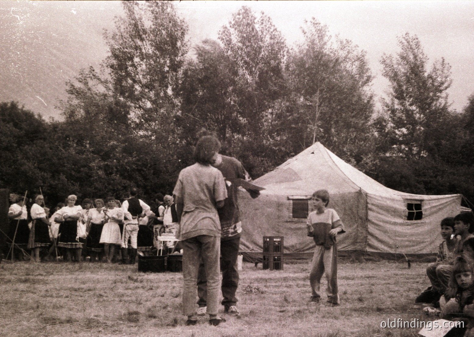 Vintage black-and-white photo of a group camping event in a grassy clearing. Central tent features a red cross emblem, suggesting a scout or youth organization gathering. Adults and children in casual 1960s-70s attire—bell-bottoms, button-ups, and headscarves—engage in activities. One man in a striped shirt points toward the tent, while others sit or stand around a small fire pit. Forest backdrop enhances the outdoor setting.