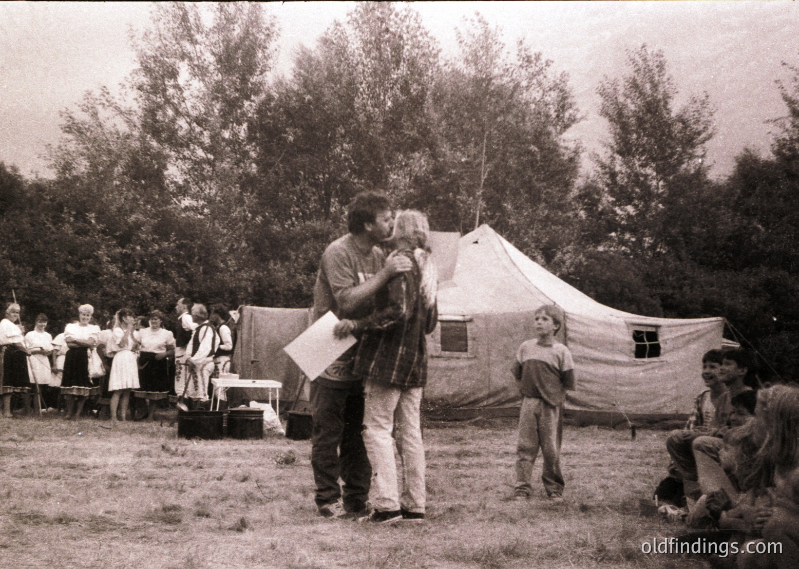 Mid-20th century outdoor gathering at a makeshift campsite with canvas tents. A man in a striped shirt embraces a child while holding a paper, surrounded by seated adults and children in casual attire. Wooden chairs and a table suggest a communal meal or event. Lush greenery and trees frame the scene, indicating a rural or park setting. Likely