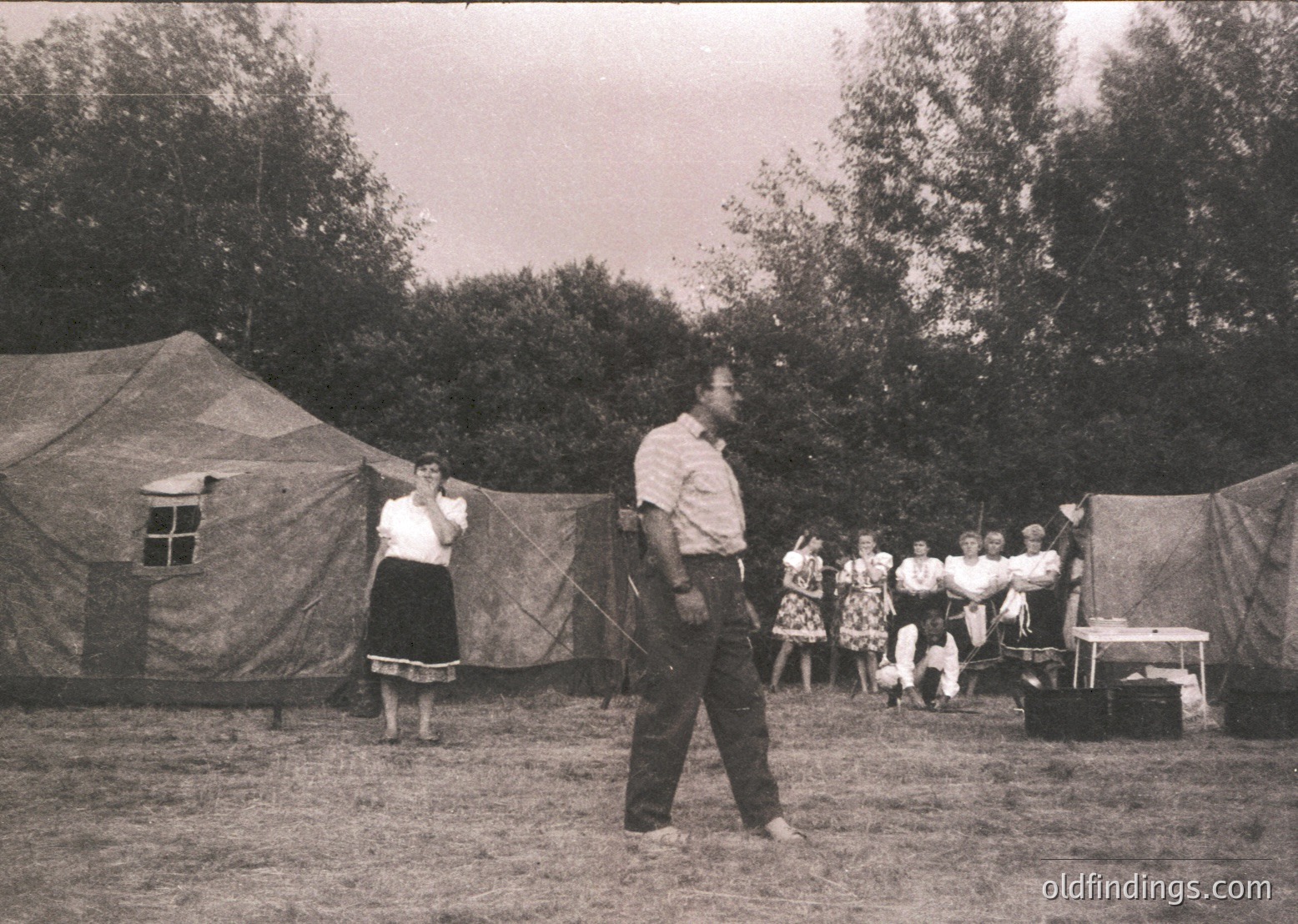 Mid-20th century outdoor gathering in a tented camp setting, likely 1950s–1960s. A man in a white shirt and dark pants performs a traditional dance while others observe. Women wear full skirts and blouses; men in shirts and trousers. Canvas tents and wooden crates suggest a rural or festival environment.