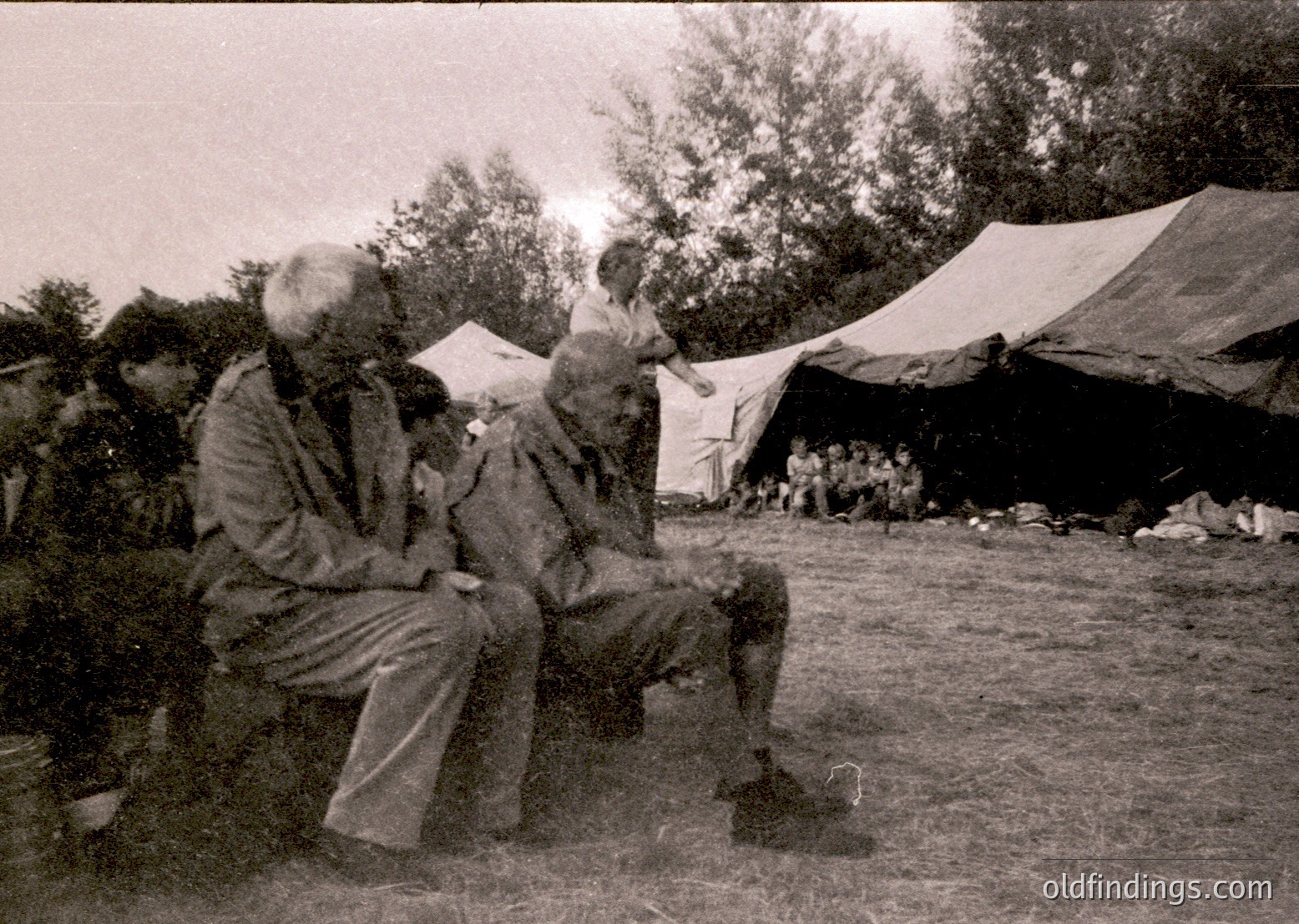 Two elderly men converse outdoors under makeshift tents in a rural setting, likely mid-20th century. Clothing suggests traditional or utilitarian styles, with one wearing a turban and both in practical workwear. Tent structures hint at a temporary camp or gathering.
