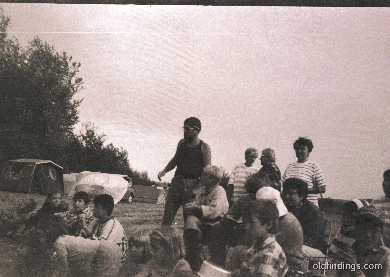 Group of people gathered outdoors in a grassy area, likely a 1970s camping scene. Central figure in sleeveless shirt poses with hands on hips. Others sit on blankets, some with backpacks and tents in background. Casual, informal gathering suggests a communal or family outing.