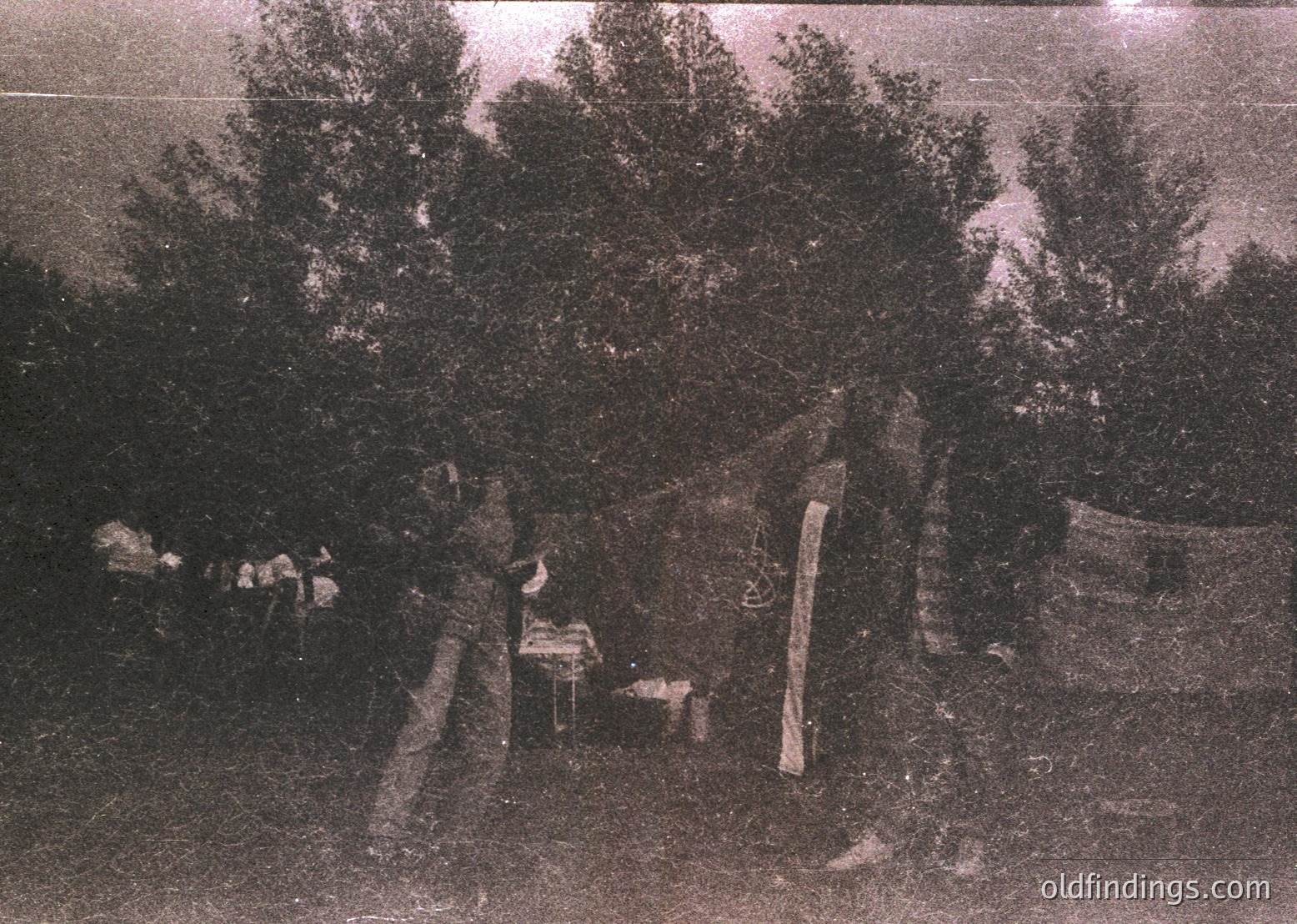 Vintage sepia-toned photo of a rural campsite with a lone figure seated near a small wooden table and chair. Surrounding foliage includes dense trees and shrubs, suggesting a forested area. Tent poles and fabric remnants indicate temporary shelter. Likely late 19th to early 20th century, outdoor adventure or exploration context.
