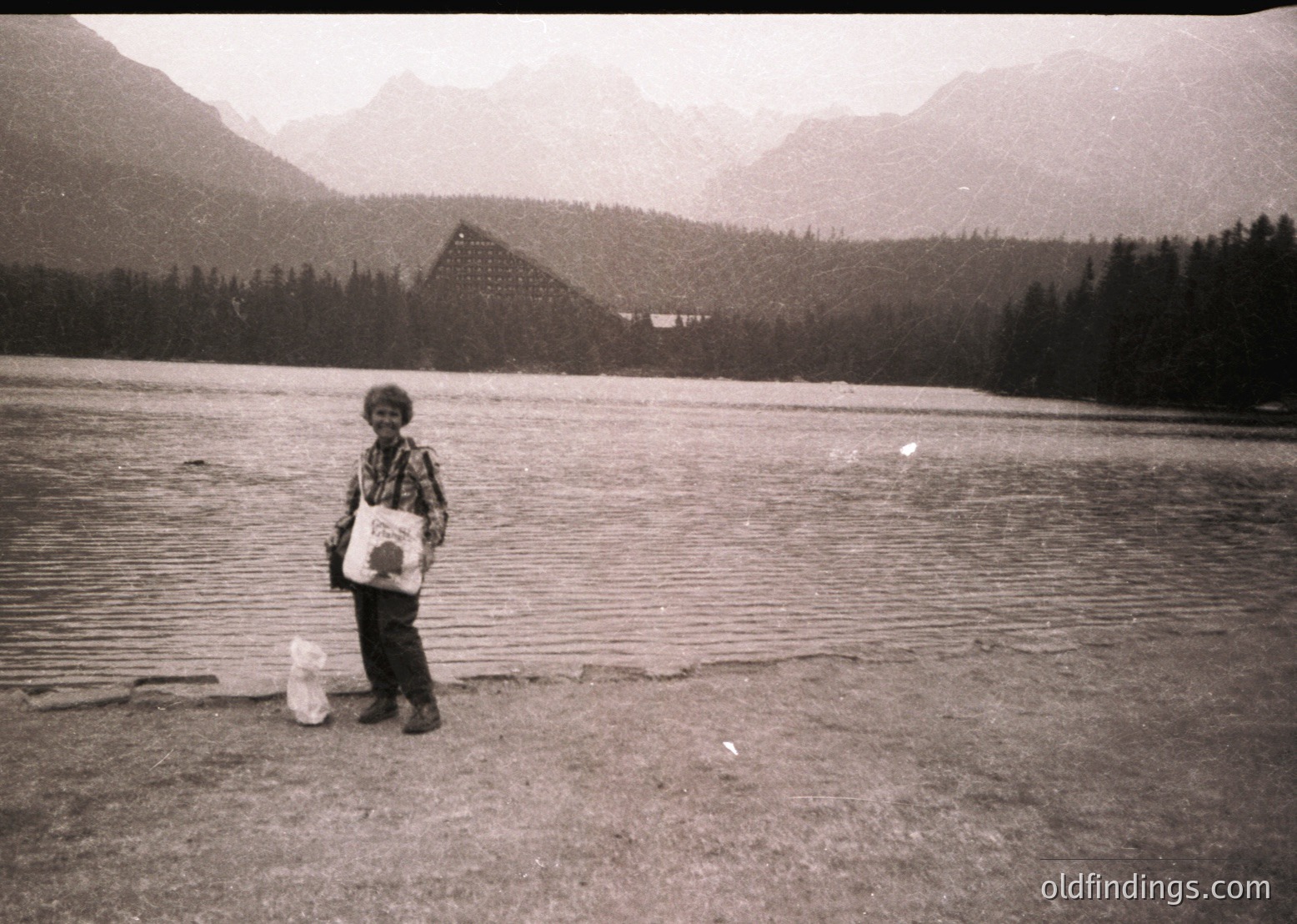 Child stands by a serene alpine lake, holding a white plastic bag and a cylindrical container, likely for fishing or collecting water. Snow-capped peaks and dense forest frame the scene, with a pyramid-shaped structure visible in the background. Black-and-white, mid-20th century (1950s–1970s) mountain photography.