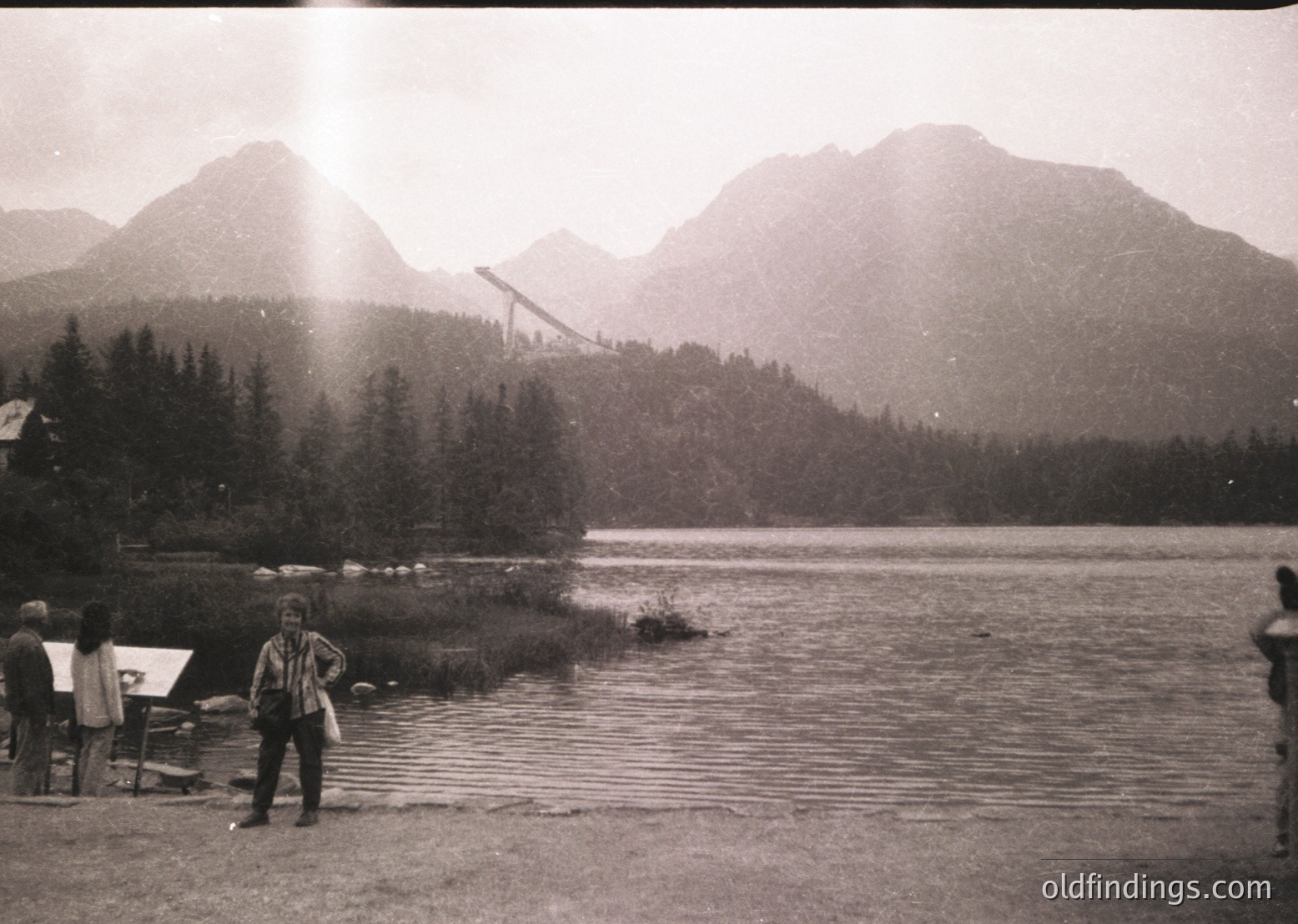 Black-and-white shot of a mountainous lakeside scene with a suspension bridge in mid-construction, surrounded by dense forest. Two figures stand near the water’s edge, one holding a bag. Mid-20th century alpine tourism vibe.