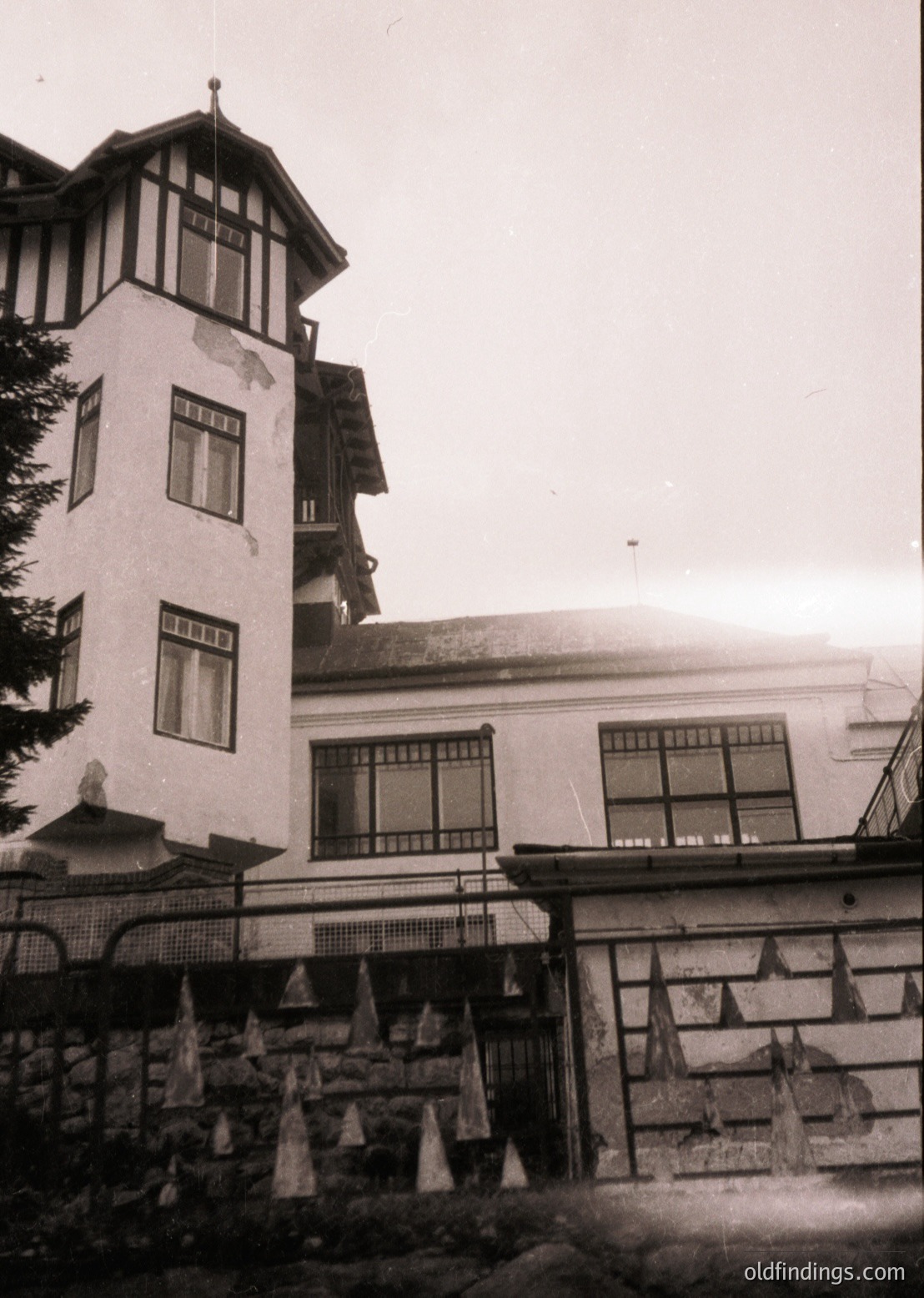 Vintage alpine resort building with Art Nouveau influences: multi-story structure featuring a corner tower with decorative brackets, large arched windows, and a sloped roof. Stone foundation with wrought-iron railings. Likely mid-20th century mountain lodge.