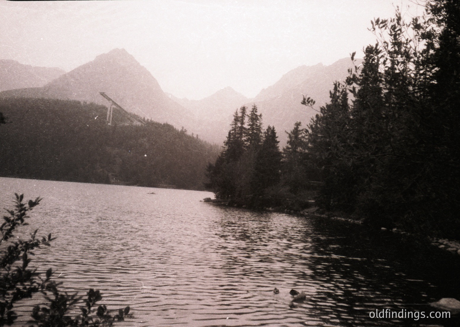 Black-and-white alpine lake framed by dense coniferous forest and rugged peaks, likely mid-20th century. Distant ski lift cables suggest early mountain tourism development. Calm waters reflect muted light, emphasizing serene natural beauty.