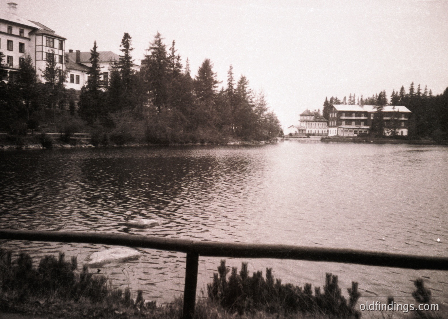 Vintage black-and-white lakeside resort with two-story wooden buildings surrounded by dense pine forest. Reflective water and wooden railing in foreground suggest early 20th-century alpine tourism.
