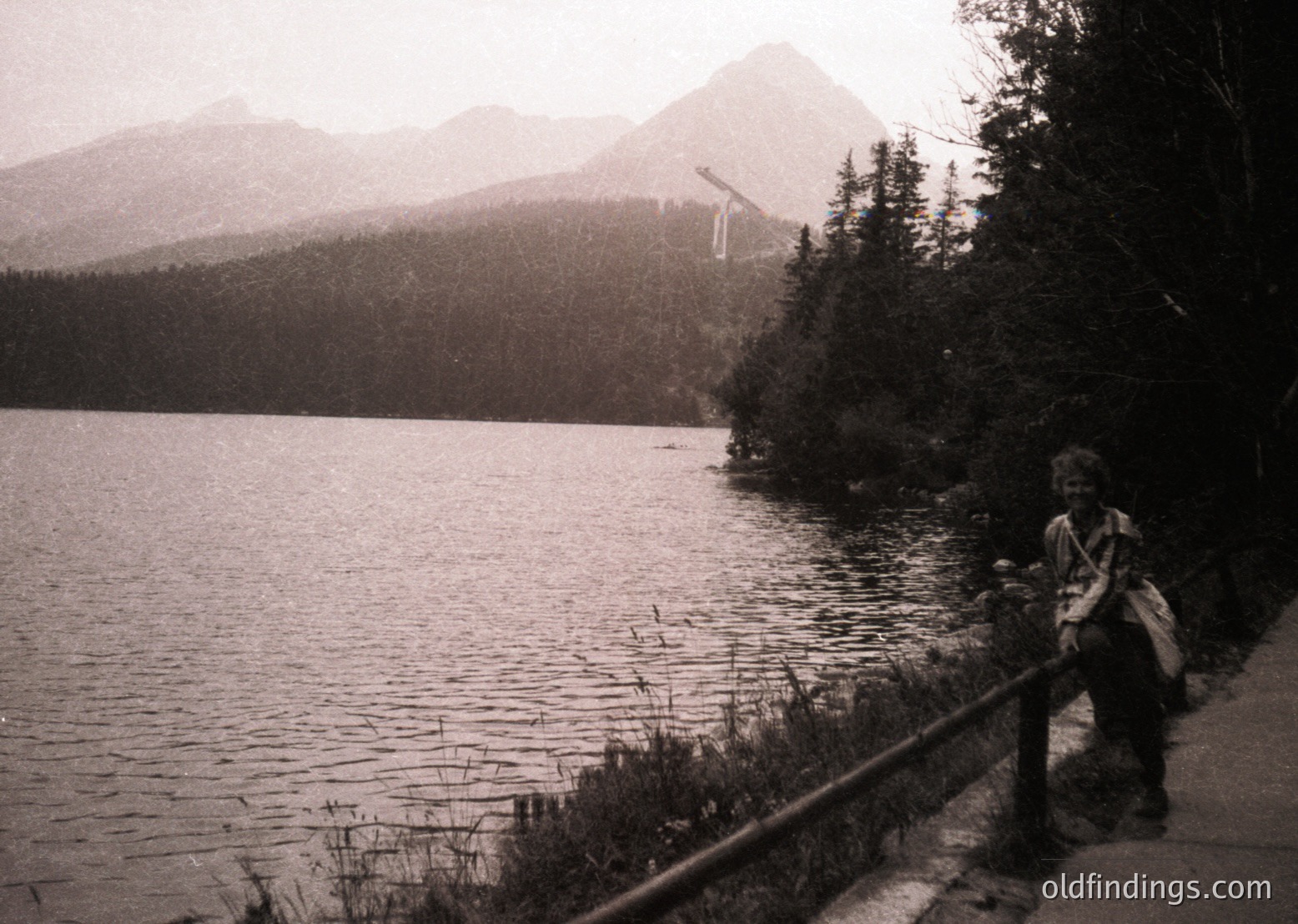 Black-and-white shot of a lone figure standing on a lakeside wooden path, surrounded by dense coniferous forest and towering alpine peaks. The person holds a camera, suggesting mid-20th century travel photography. Likely --- *Note: Exact location indeterminable, but stylistic cues align with European alpine regions.*
