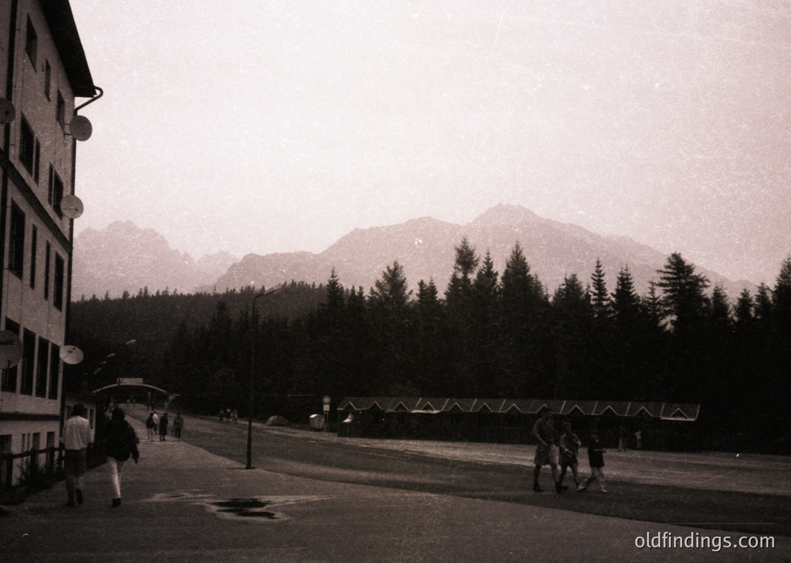 Mid-century alpine resort scene with multi-story concrete building and forested mountain backdrop. Snow-capped peaks frame the horizon under overcast skies. Pedestrians in winter attire walk along a wet, paved street. Decorative holiday lights strung across the road suggest festive season.