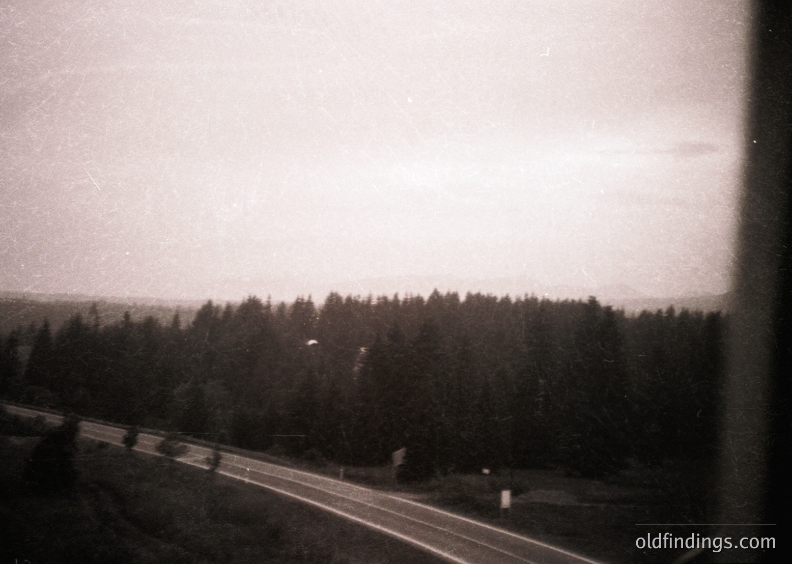 Vintage black-and-white shot of a rural road bordered by dense evergreen forest under overcast skies. The composition suggests a mid-20th century travel scene, likely or . The road’s narrowness and lack of modern infrastructure hint at a remote or less-developed area.