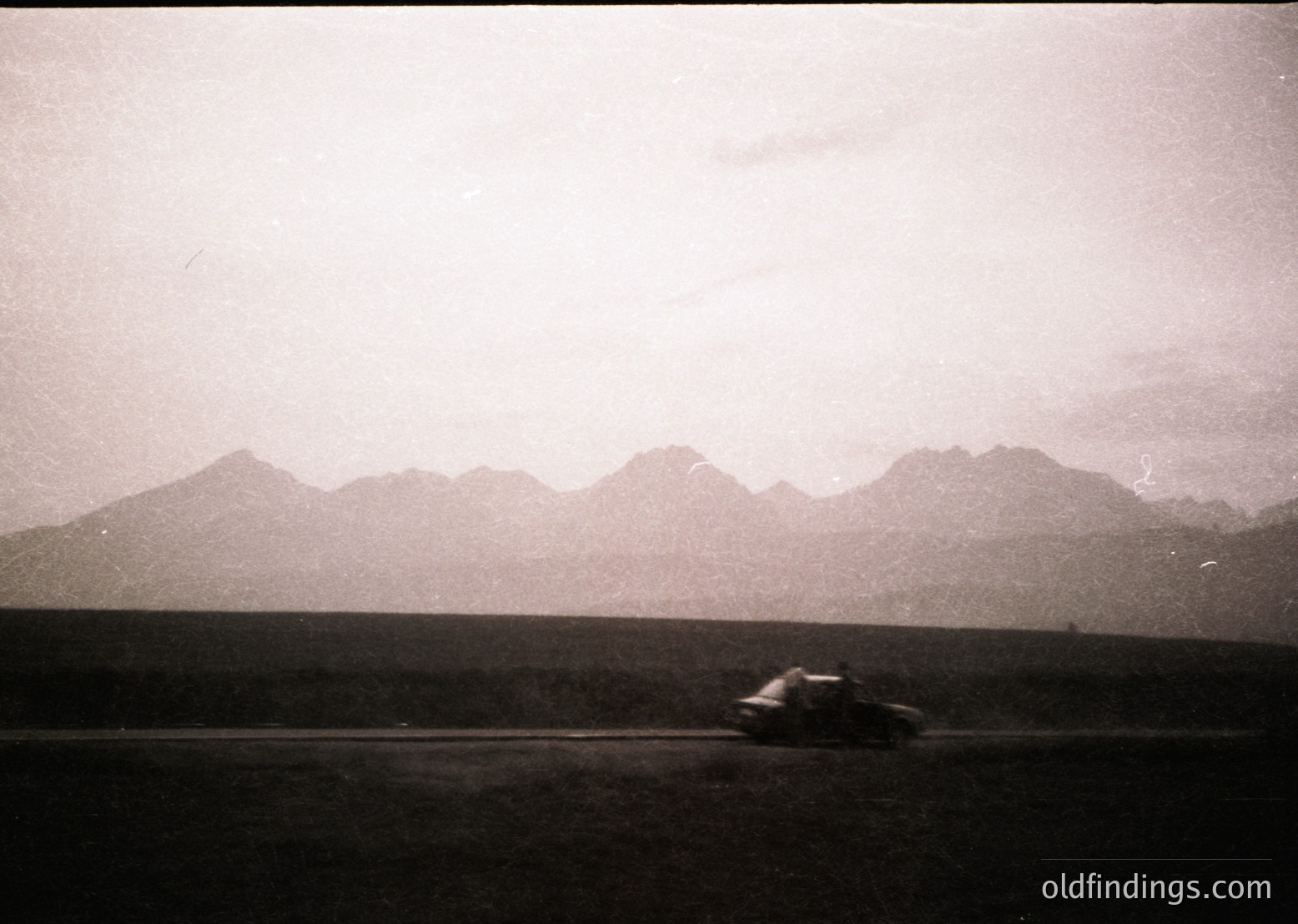 Vintage black-and-white shot of a lone vintage car driving on a winding rural road, flanked by misty mountain ranges. The sepia tone suggests mid-20th century (1950s–1960s). Ideal for nostalgic travel or automotive design references.
