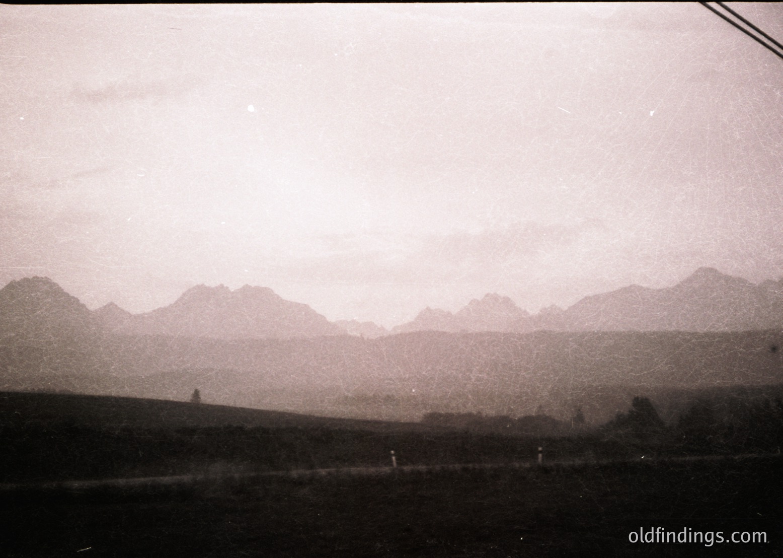 Vintage sepia-toned landscape featuring jagged mountain range under overcast skies. Foreground shows sparse vegetation and a faint road or path. Likely early 20th-century European alpine scenery.