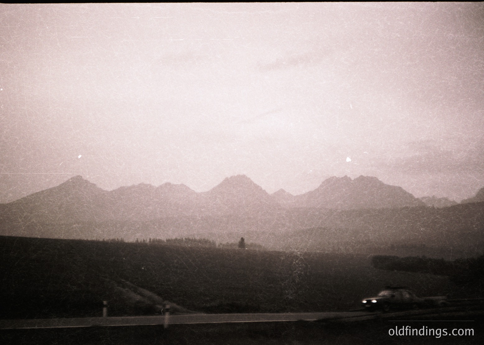 Vintage black-and-white shot of a lone car driving along a winding alpine road, flanked by dense conifer forests and jagged peaks. Fog obscures mid-ground, enhancing misty atmosphere. Likely 1950s–1970s European or North American mountain region.