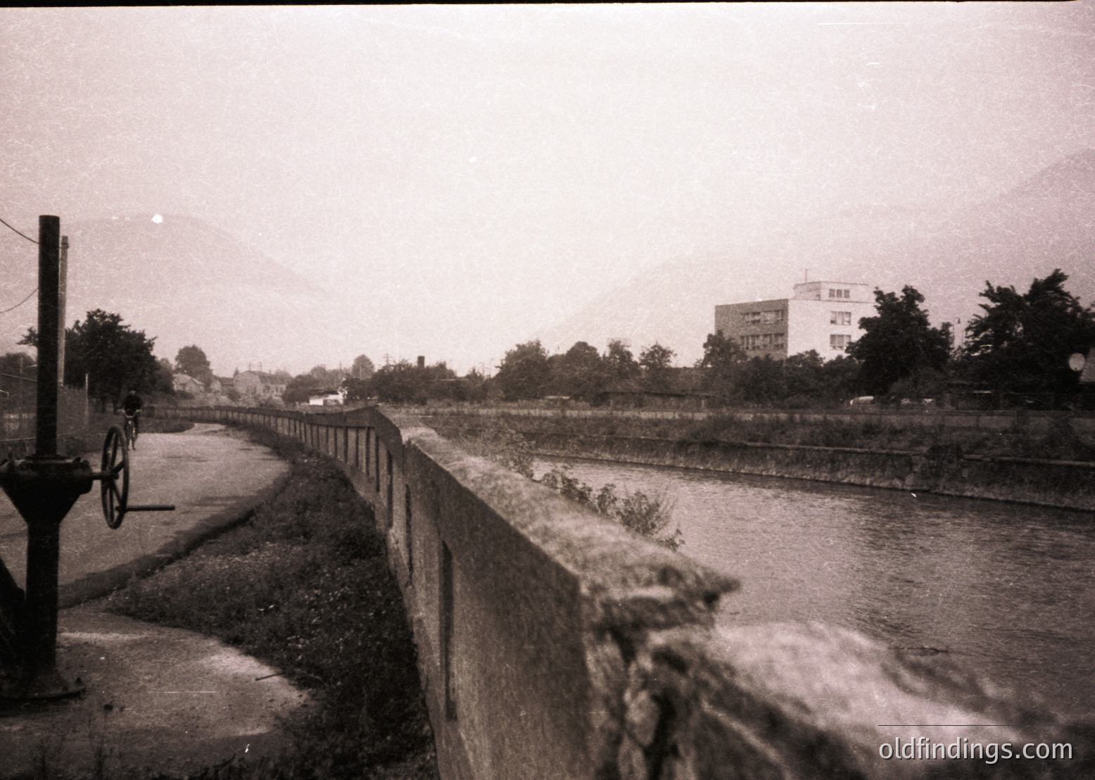 Black-and-white urban canal scene with concrete embankment and rustic fence. A lone cyclist rides along a narrow road beside the water, flanked by sparse vegetation and distant mid-rise buildings. Overcast skies enhance the vintage atmosphere. Likely 1960s–1980s European industrial city.