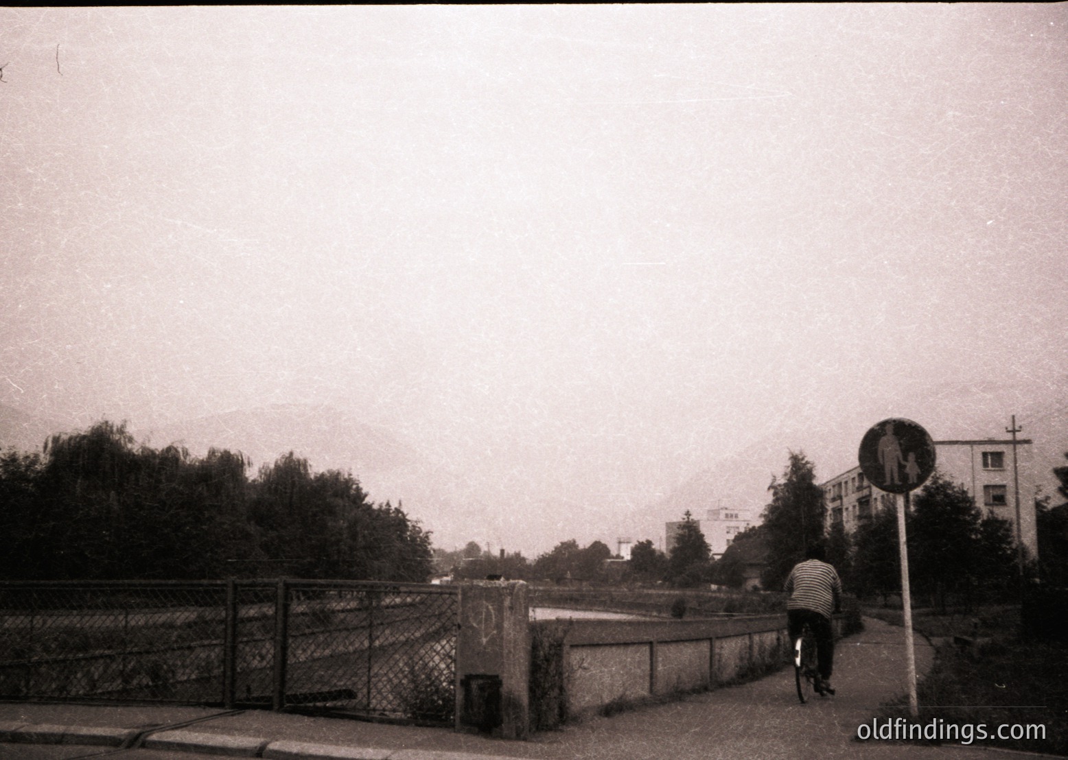 Black-and-white street scene featuring a lone cyclist crossing a rusted metal bridge over a body of water. Surrounding greenery and mid-rise residential buildings in background. Graffiti on bridge railing. Vintage urban aesthetic, likely 1970s–1990s.