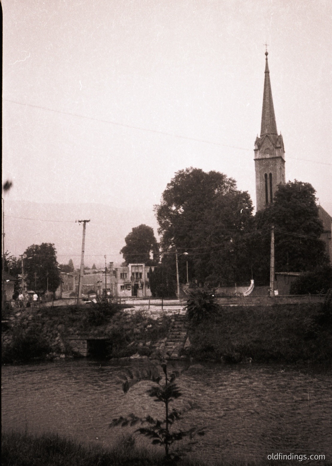 Black-and-white street scene featuring a Gothic Revival church spire with pointed arches, likely 19th-century European architecture. Stone bridge and riverbank with minimal vegetation frame the foreground. Overhead wires and sparse urban structures suggest early 20th-century town planning.