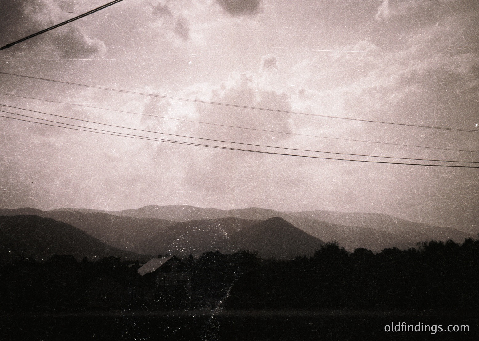 Black-and-white shot of a rural landscape framed by power lines. Dense forest and rolling hills dominate the midground, while a full moon casts dramatic light on clouds and terrain. Likely 20th-century vintage, evoking mid-century travel or documentary photography.