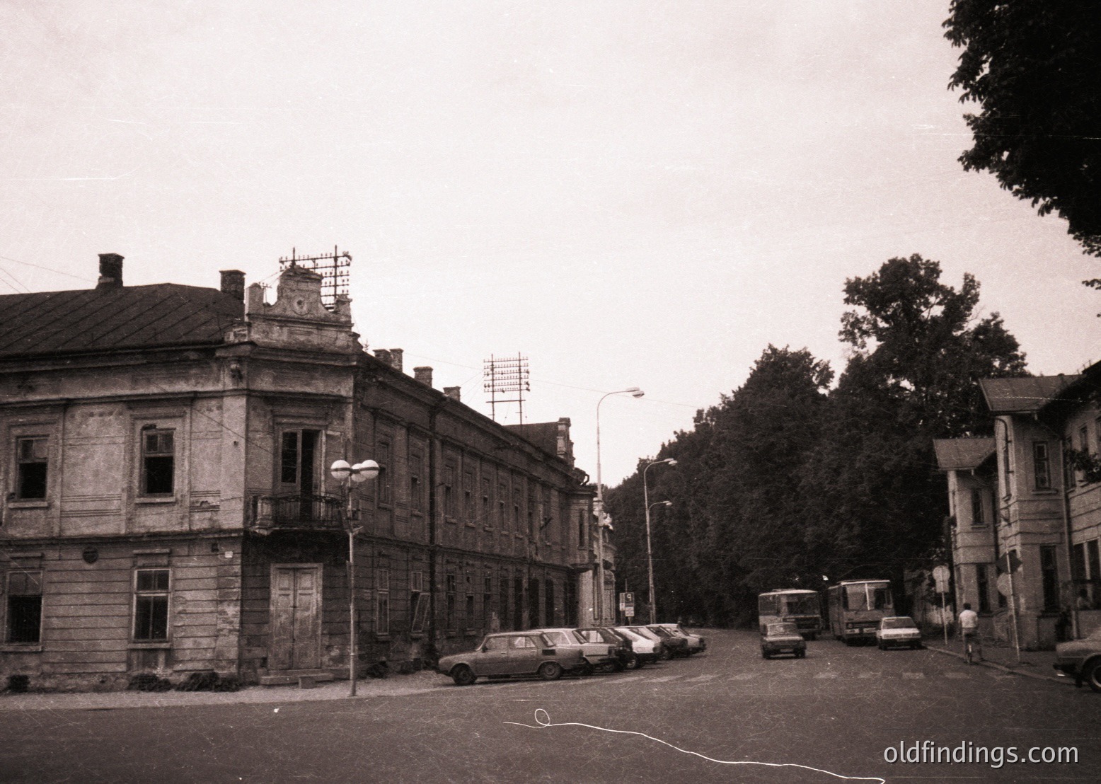 Vintage black-and-white street scene featuring Soviet-era architecture with a two-story stone building showcasing classical details like arched windows and a decorative cornice. Parked cars (likely Ladas) and a bus suggest mid-20th-century urban life. Overhead power lines and sparse greenery indicate an older, possibly Eastern European cityscape.