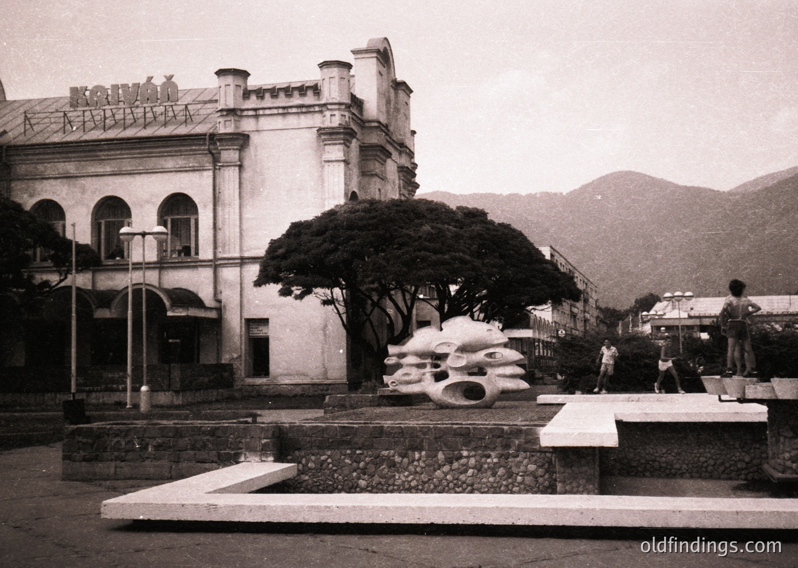 Neoclassical building with arched windows and decorative roofline in a seaside plaza, mid-20th century. Abstract concrete wave sculpture in foreground, surrounded by stone steps and lampposts. Mountain backdrop suggests coastal urban setting.
