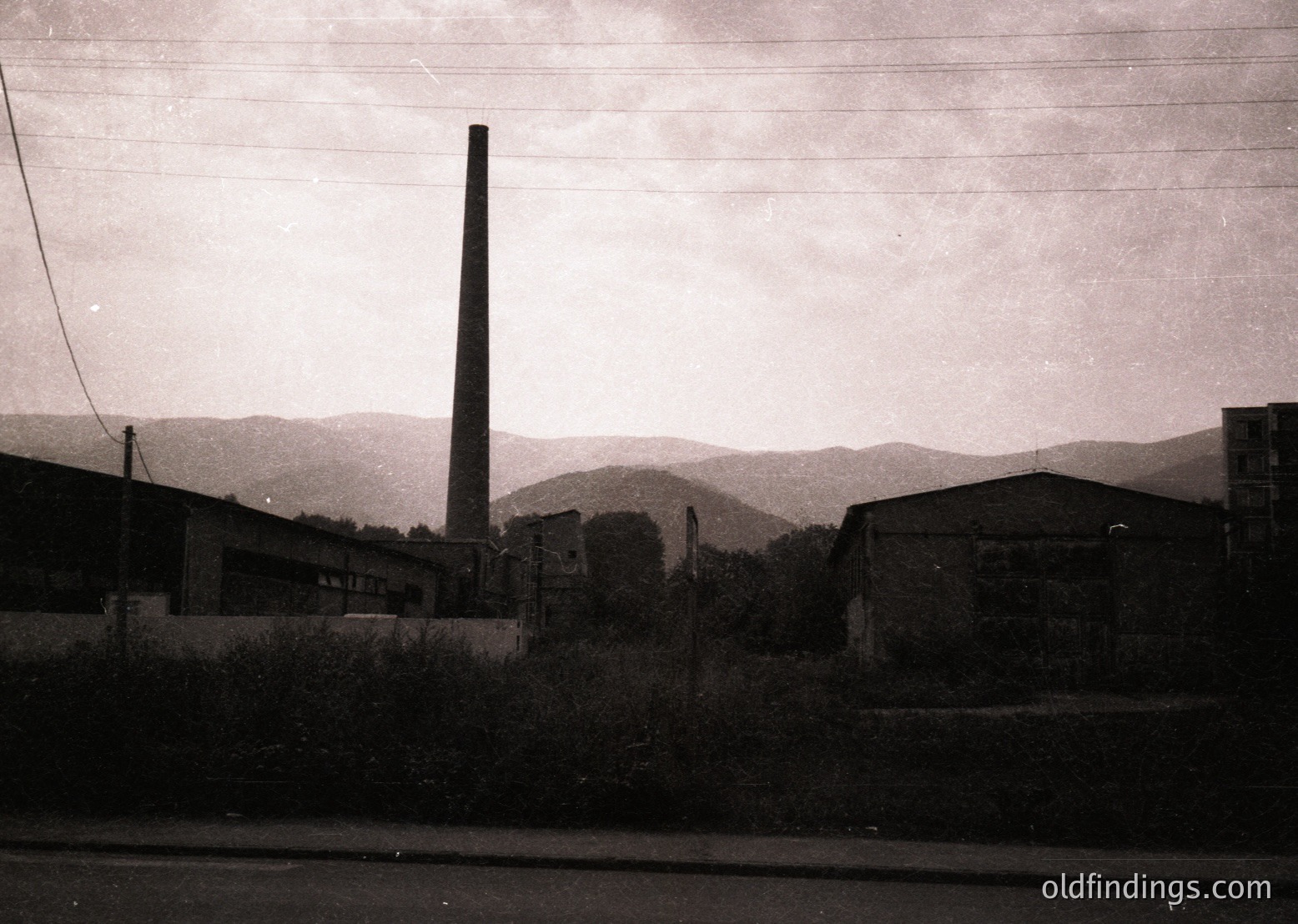 Black-and-white industrial landscape featuring a tall brick smokestack and adjacent brick buildings, likely a factory or power plant. Overgrown vegetation and a rural road frame the scene, with rolling hills in the background. Style suggests mid-20th century (1950s–1970s) industrial architecture.