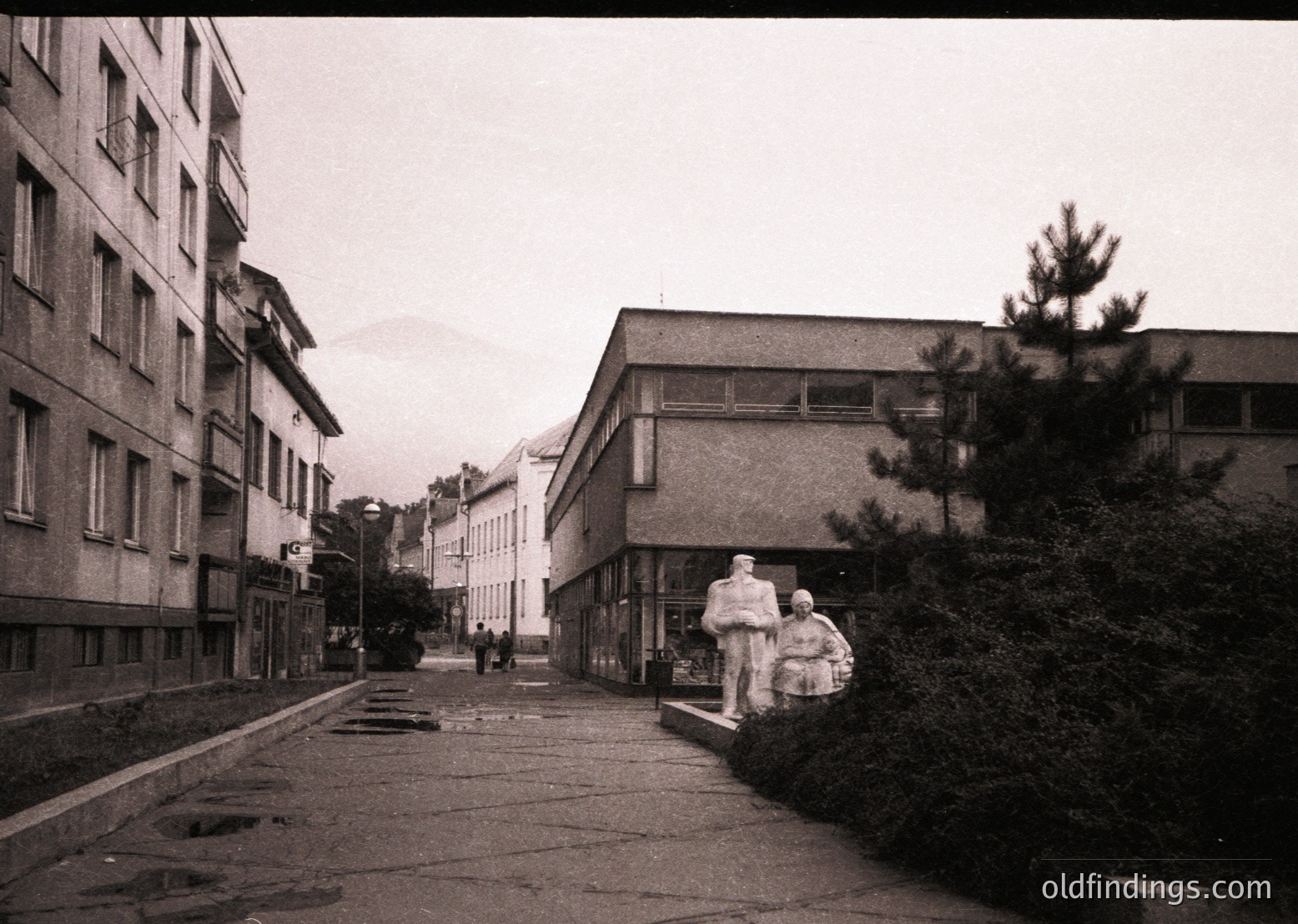 Mid-century urban street with Soviet-era Brutalist architecture—concrete apartment blocks and low-rise institutional buildings lining a paved walkway. Two statuesque figures in dynamic poses flank a stairway entrance. Distant mountain silhouette suggests alpine setting. Likely Eastern Bloc, 1960s–70s.
