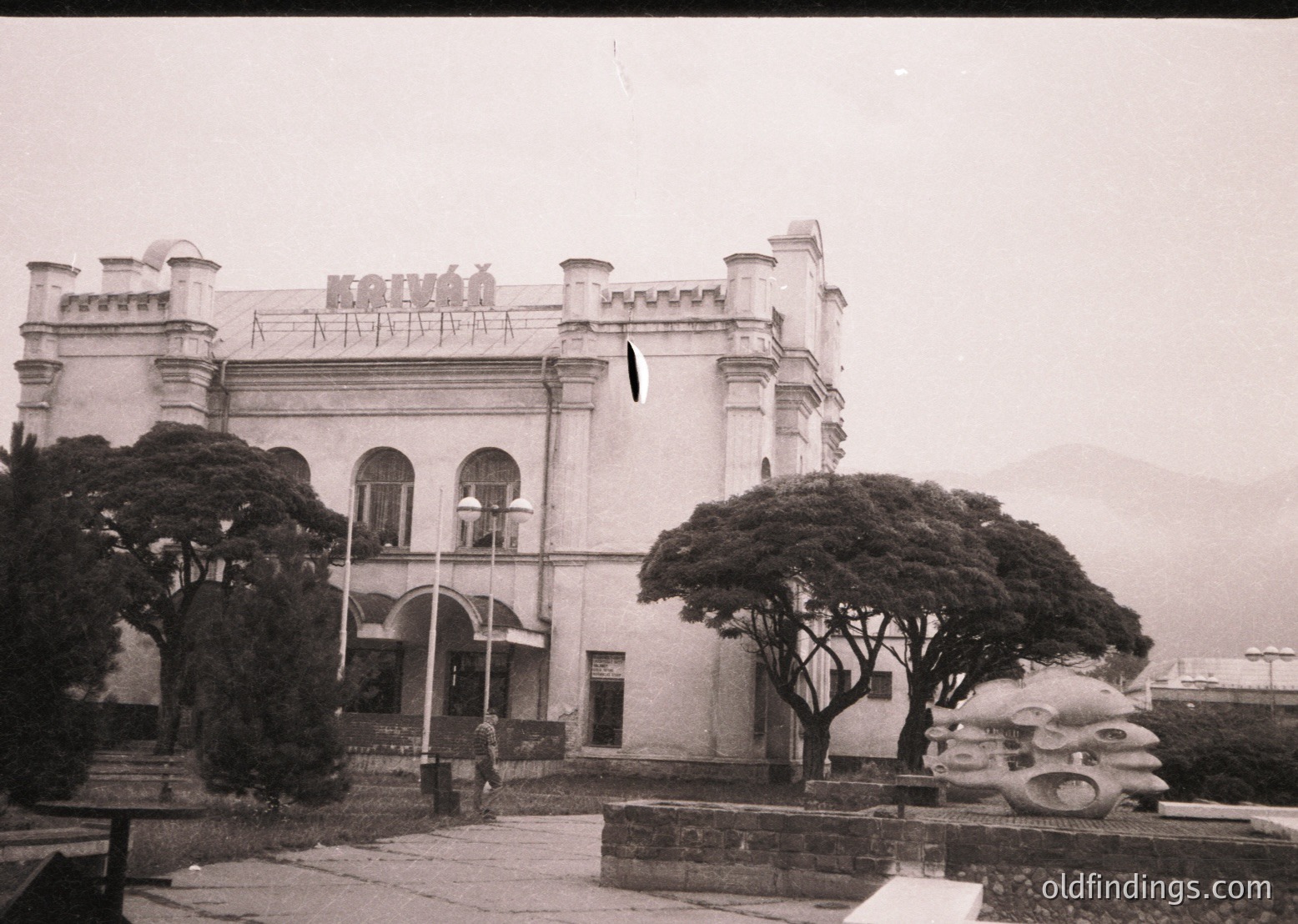 Neoclassical seaside villa with crenellated roofline, arched windows, and symmetrical towers—likely a Bulgarian Black Sea resort from the early 20th century. Foggy coastal backdrop hints at Varna or Sozopol. Decorative stone balustrade and vintage car tires repurposed as planters.