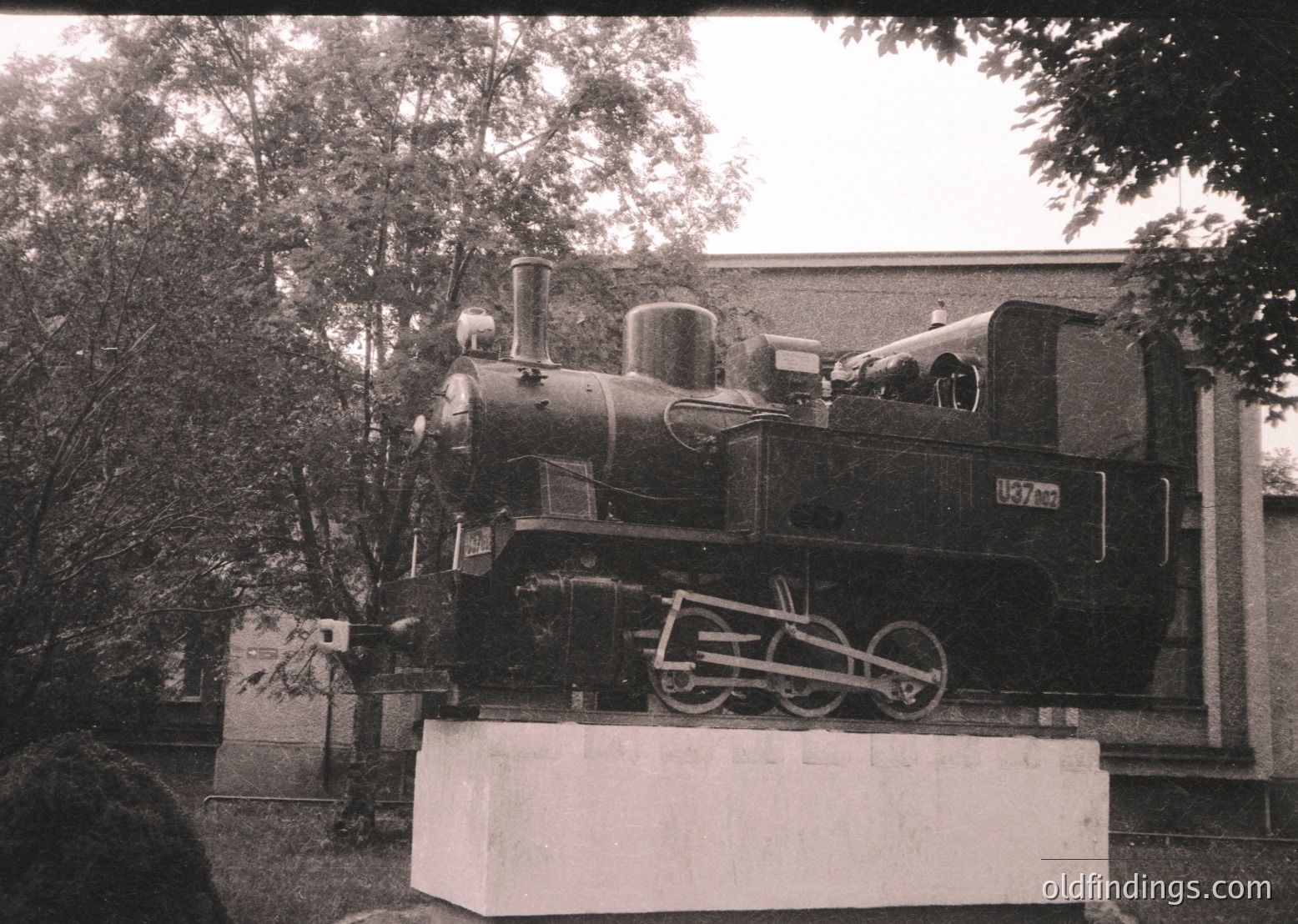 Vintage steam locomotive mounted on a concrete pedestal, likely a preserved exhibit. Visible number "157" and "1908" on the engine, suggesting early 20th-century European design. Surrounding greenery and partial building structure indicate an outdoor museum or park setting.