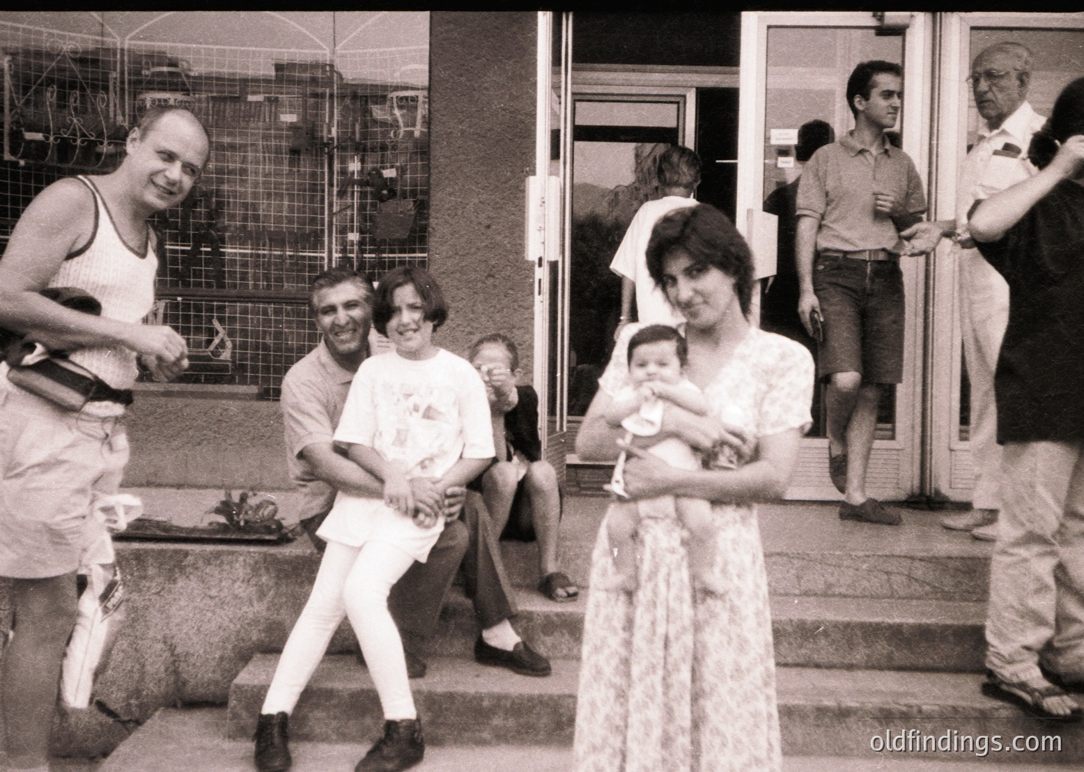 Black-and-white candid shot of a lively 1970s gathering outside a building with industrial scaffolding. Central figures: a woman in a lace dress cradling a baby, a man in a tank top mid-laugh, and a seated group in casual summer attire. Urban setting suggests a communal or celebratory event.