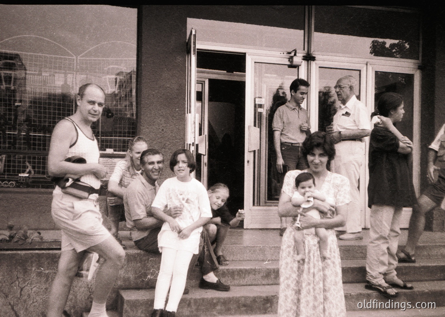 A candid 1970s-era black-and-white photo captures a lively gathering outside a Soviet-era apartment building. A diverse group—men in tank tops/slacks, women in dresses/blouses—stands on concrete steps and porch, with one woman holding a baby. A man in a sleeveless vest poses mid-stride, while others lean or chat. A child sits on the steps, and a dog rests on the ground. The building’s utilitarian design and metal mesh balcony reflect mid-century socialist architecture.