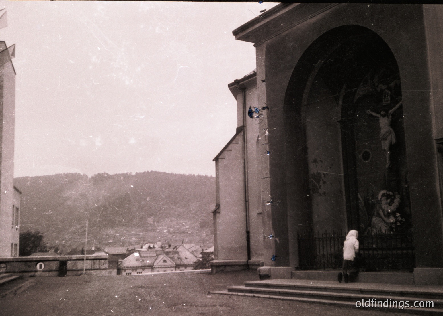 Black-and-white street scene featuring a weathered religious archway with a crucifix and saint mural. A lone figure in a long coat stands on a stone platform. Mid-20th century European village with hilly backdrop.