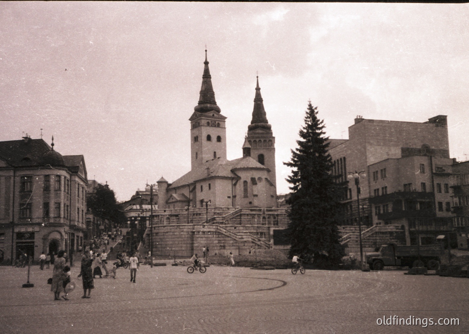 Mid-20th century street scene featuring a prominent church with twin spires and a central tower, likely European. Pedestrians, cyclists, and a horse-drawn cart populate the cobblestone plaza. Surrounding buildings exhibit early modernist architecture with flat roofs and large windows. Decorative Christmas tree suggests late 1940s–1950s holiday season.