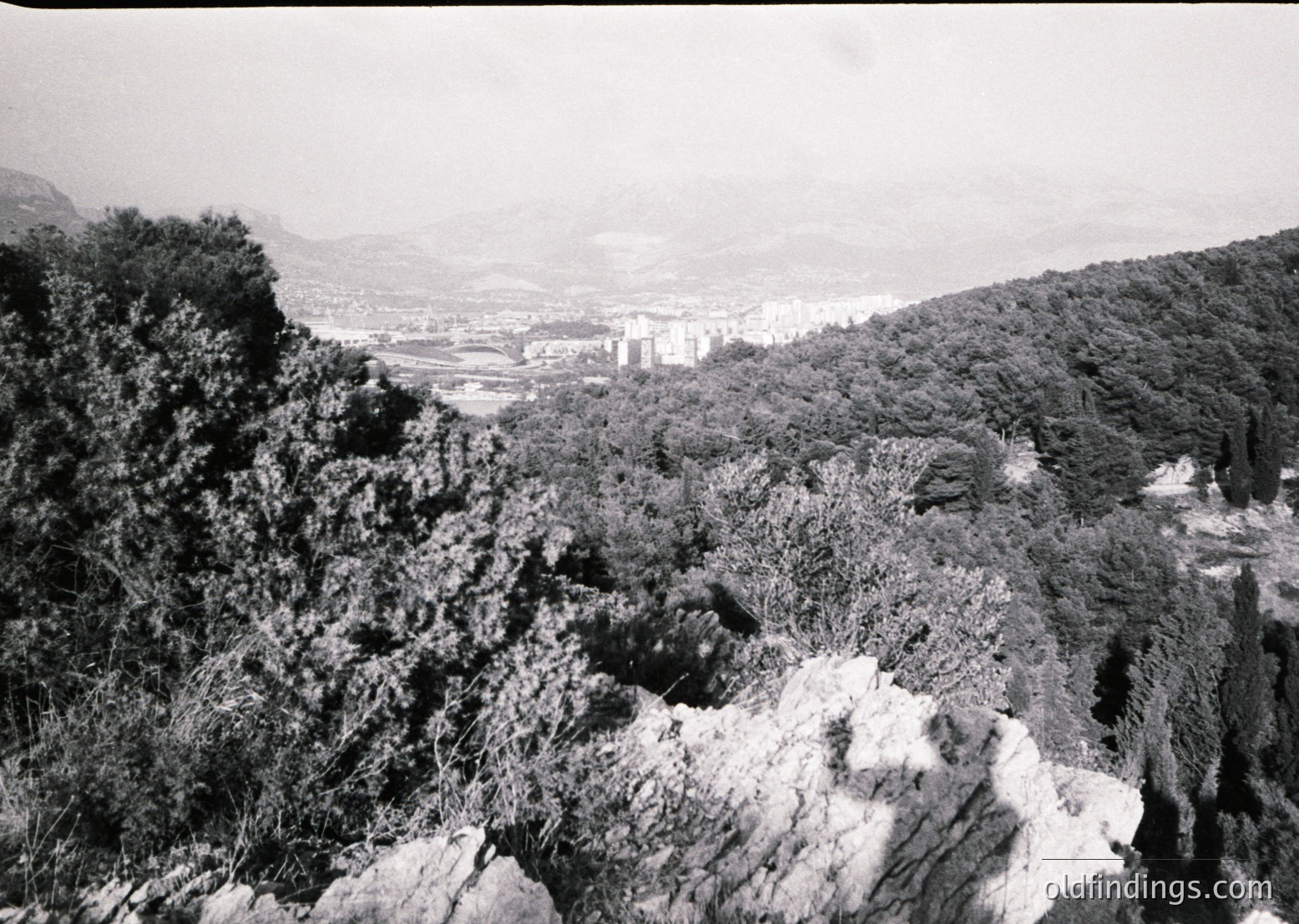 Black-and-white aerial view of rugged coastal terrain with dense forest and rocky outcrops. Distant urban sprawl and industrial structures visible along coastline, suggesting port or harbor activity. Likely mid-20th century due to monochrome and composition style.