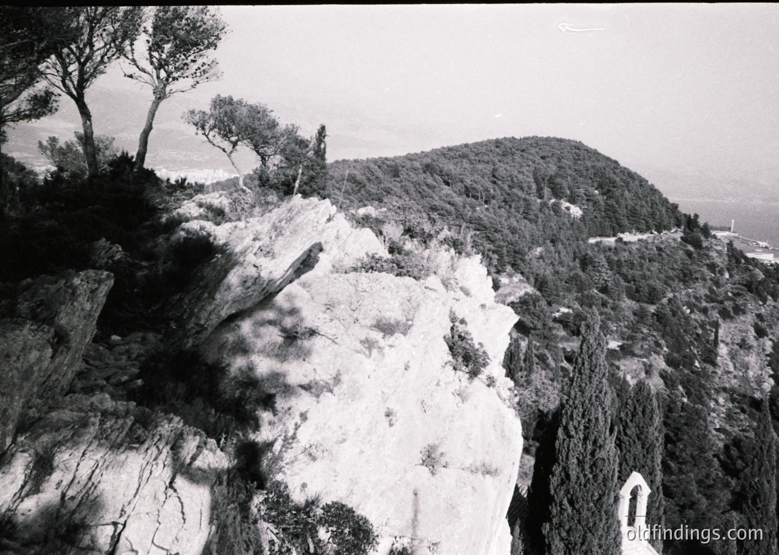 Black-and-white coastal landscape featuring jagged limestone cliffs with sparse vegetation. A lone church steeple peeks from the right, hinting at human presence. Overcast skies and misty horizon suggest a dramatic seaside setting, likely Mediterranean or Adriatic. Mid-20th century aesthetic ().