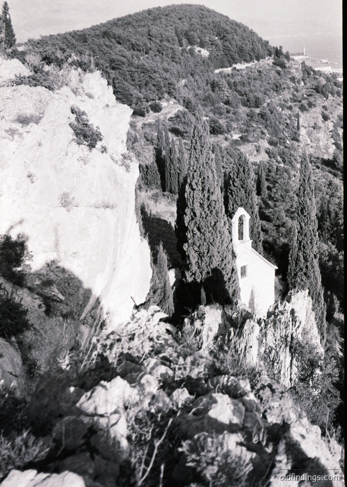 Isolated cliffside church perched on rugged terrain, surrounded by rocky slopes and sparse vegetation. Architectural details include a bell tower and arched entrance. Likely mid-20th century based on style. Dramatic composition highlights isolation and natural contrast.